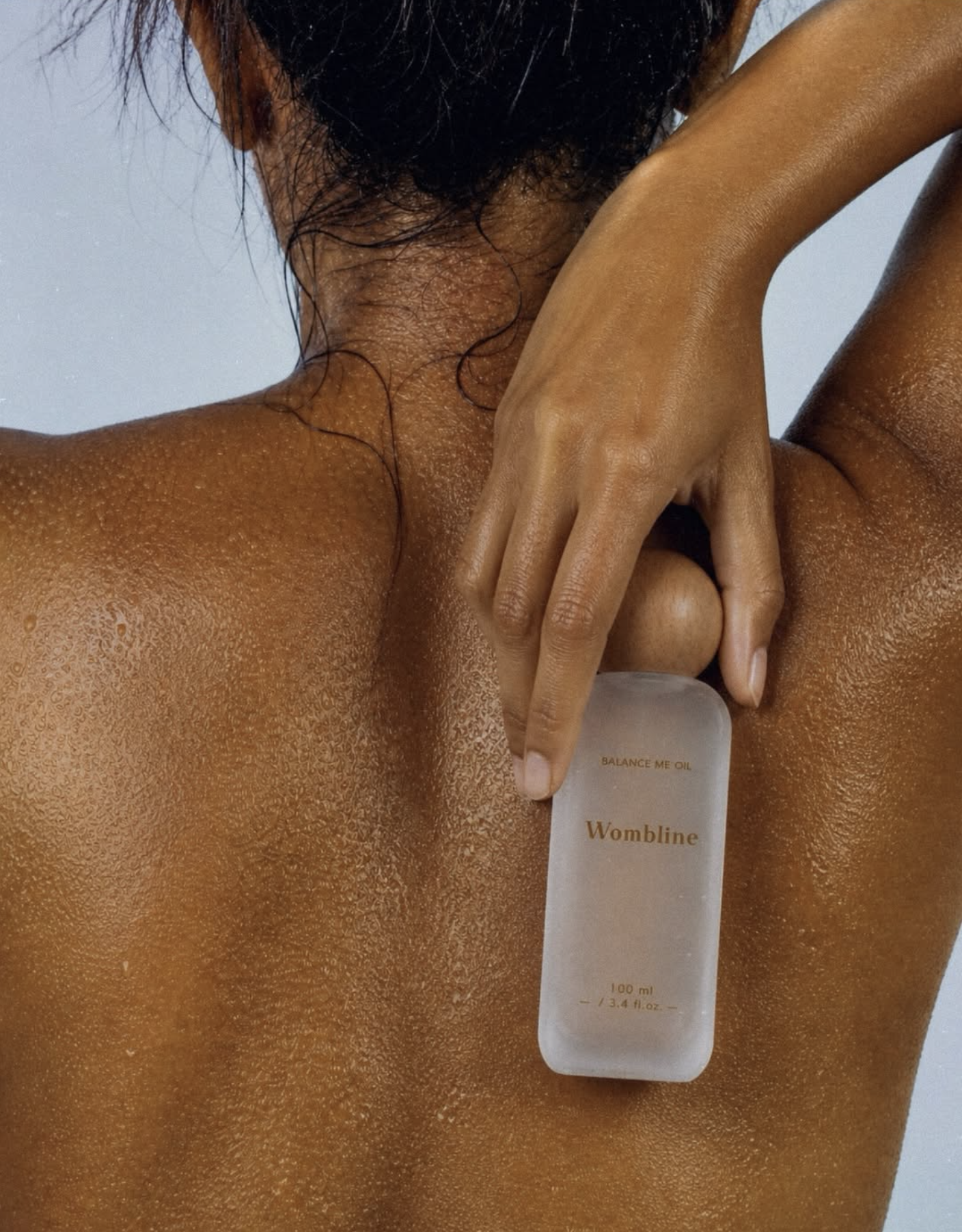 A close-up photograph of a person's back and shoulder, showing wet skin and hair, with a white bottle labeled 'Womblin' and 'Balance Me Oil' resting on their shoulder.