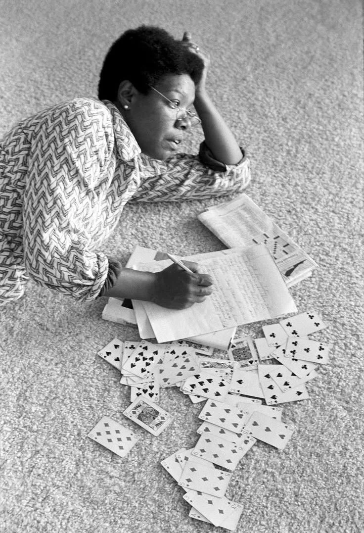 A woman lying on a carpeted floor surrounded by playing cards and writing in a notebook.