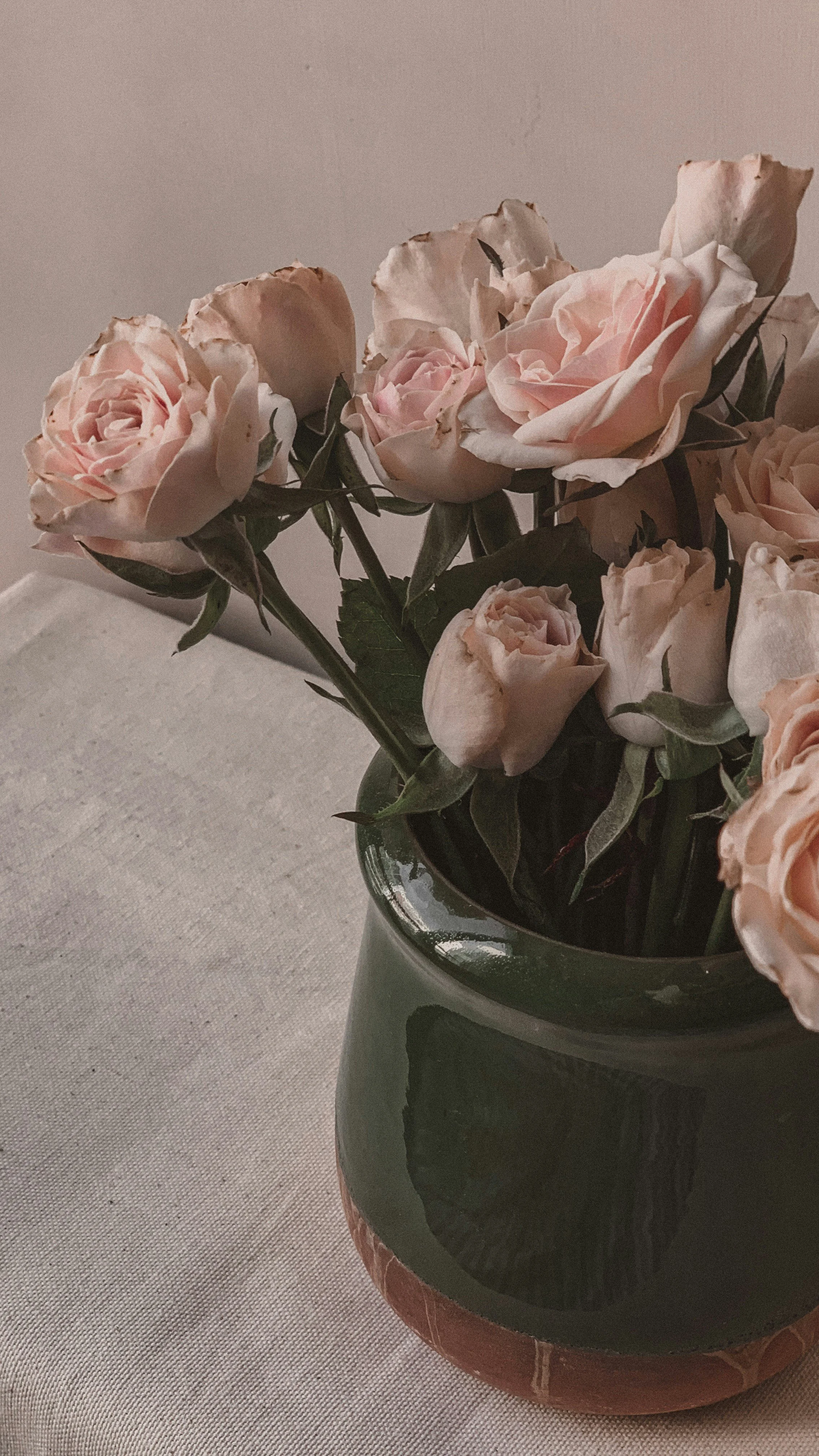 A black vase with pink roses and greenery sitting on a white textured surface against a plain background.