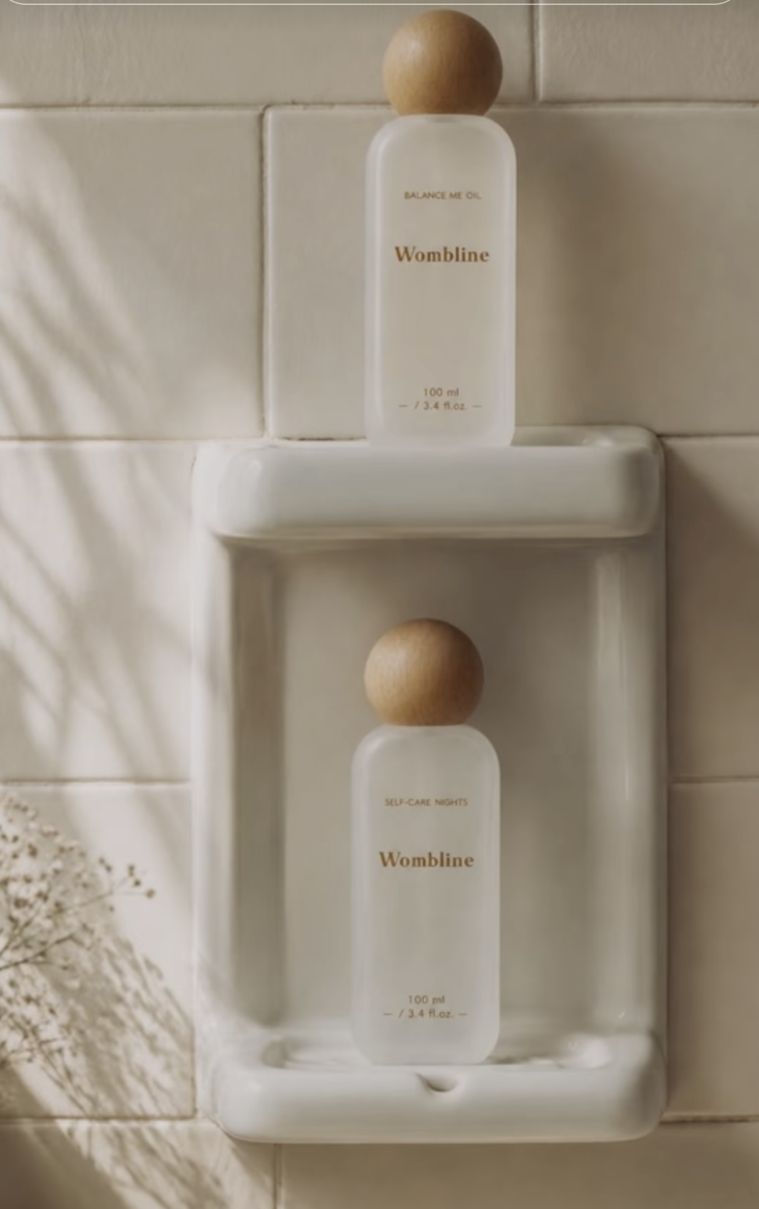 Two clear bottles with wooden caps, labeled 'Womblin' on a white corner shower shelf, with beige tiled wall background.