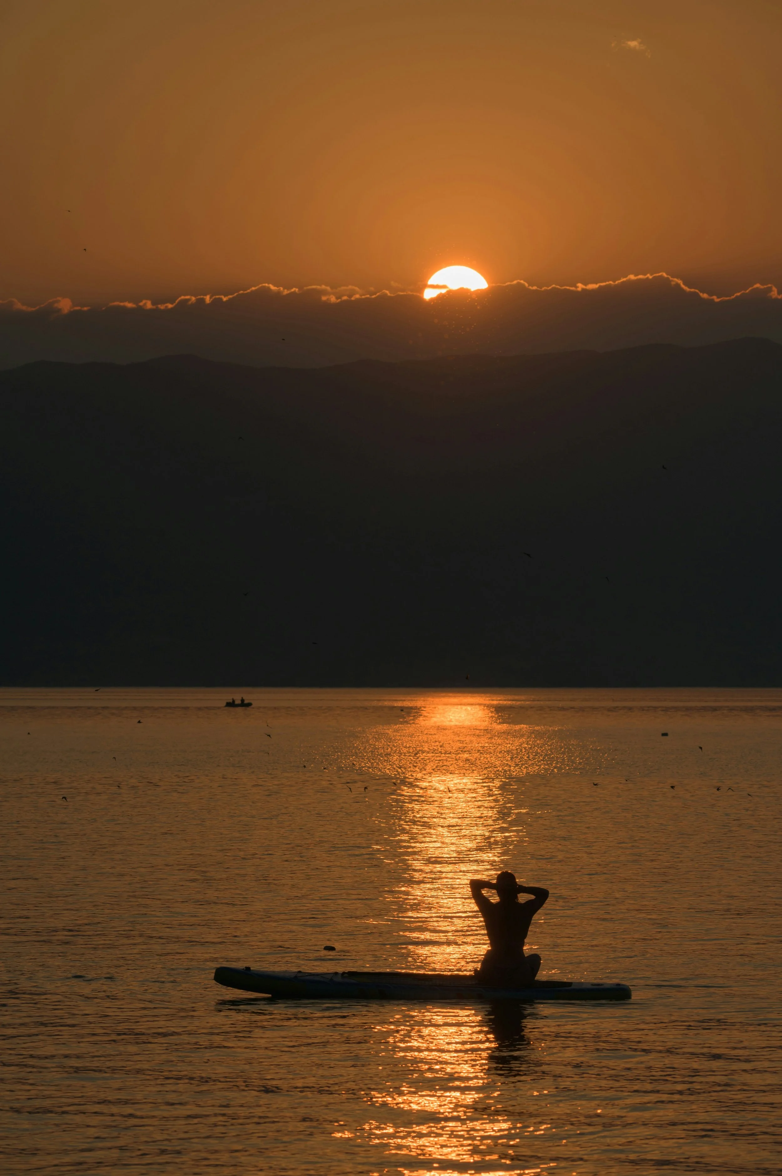 Sunset over a body of water with mountains in the background. A person is sitting on a paddleboard in the water, enjoying the view.