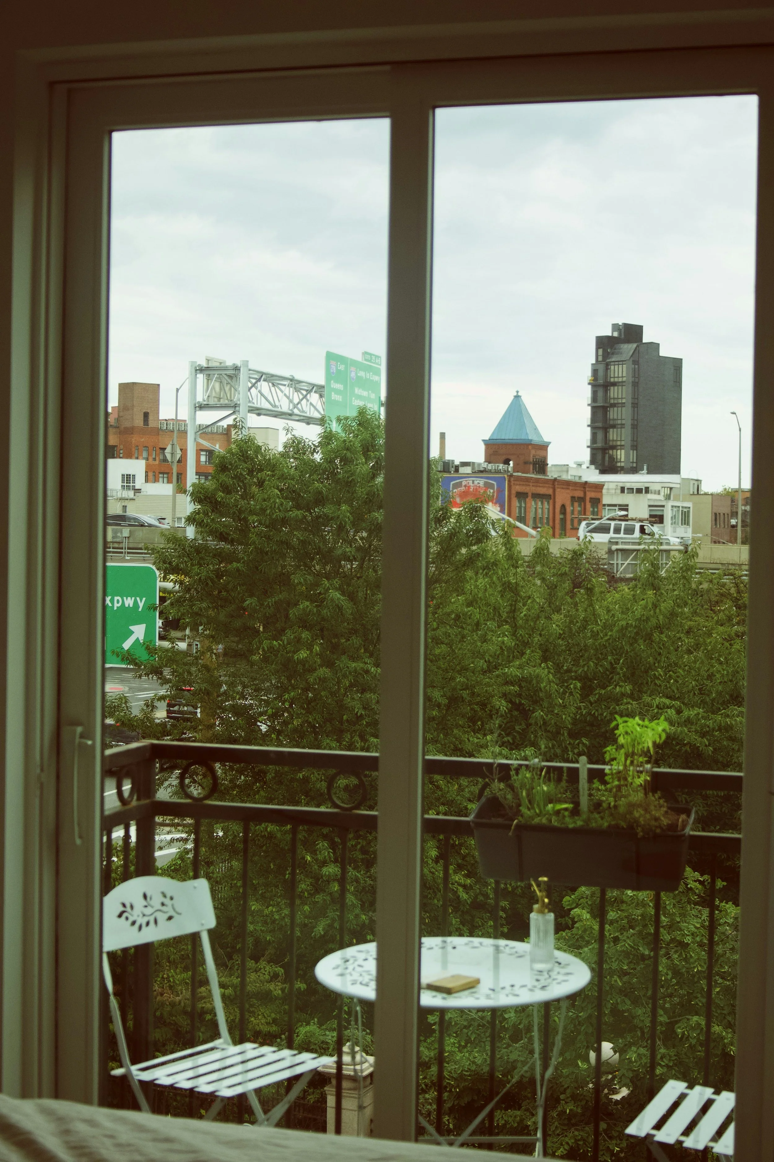 View of a cityscape through a window with a balcony, showing trees, various buildings including a tall black building, a red brick building with a pointed roof, and a highway sign, under a cloudy sky.
