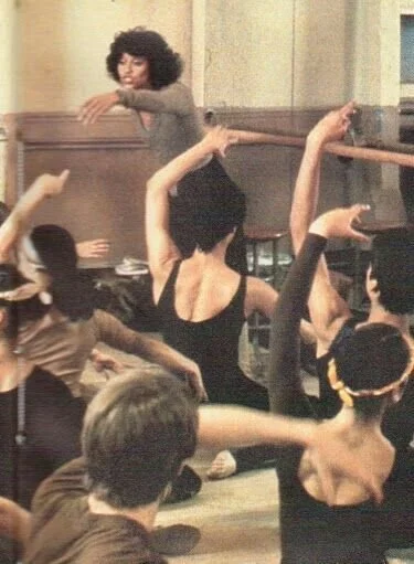 A group of dancers practicing ballet, with a woman in the background overseeing or instructing, in a studio or rehearsal space.