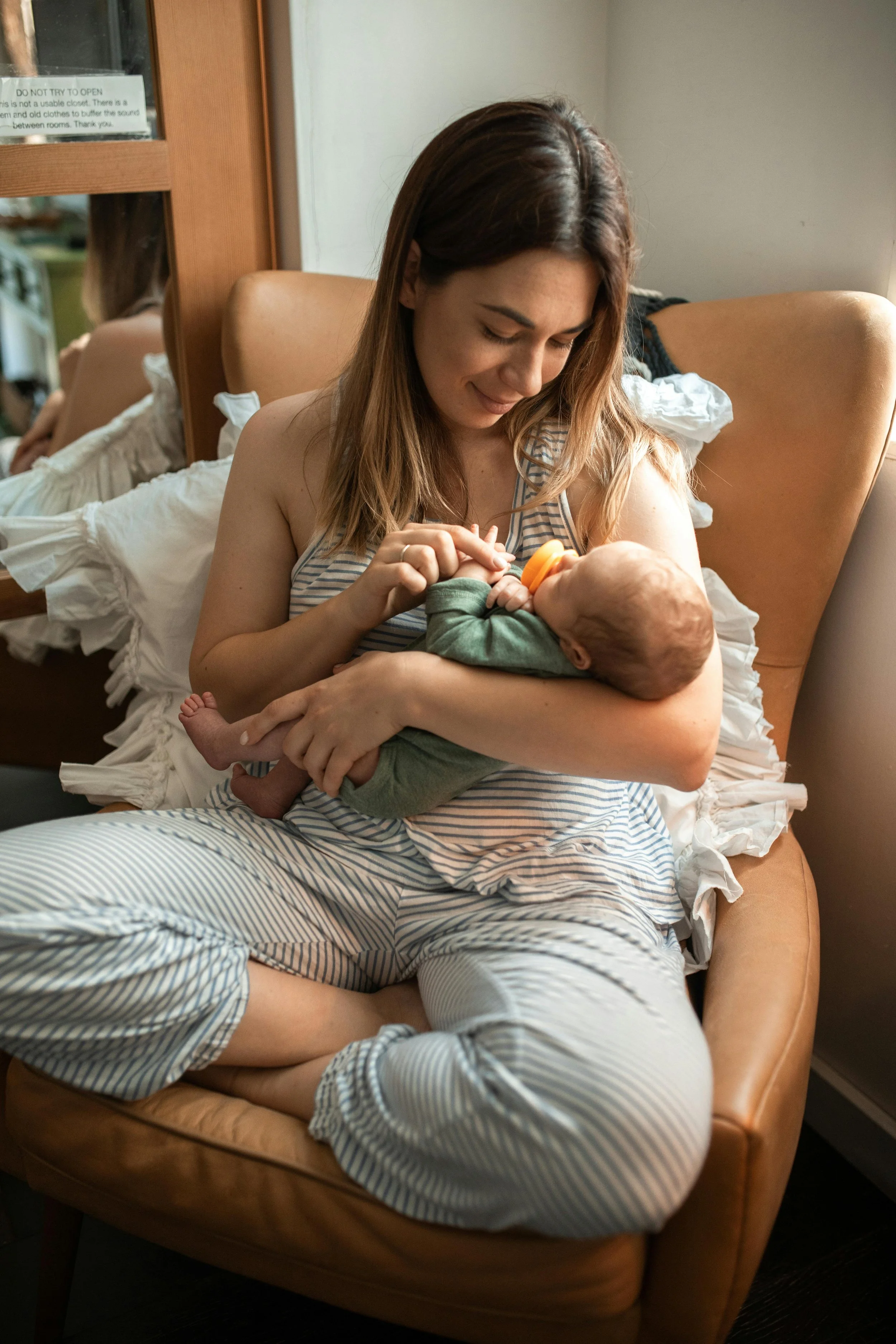 A woman sitting on a tan armchair holding a baby in her lap, feeding the baby with a bottle in a cozy, softly lit room.
