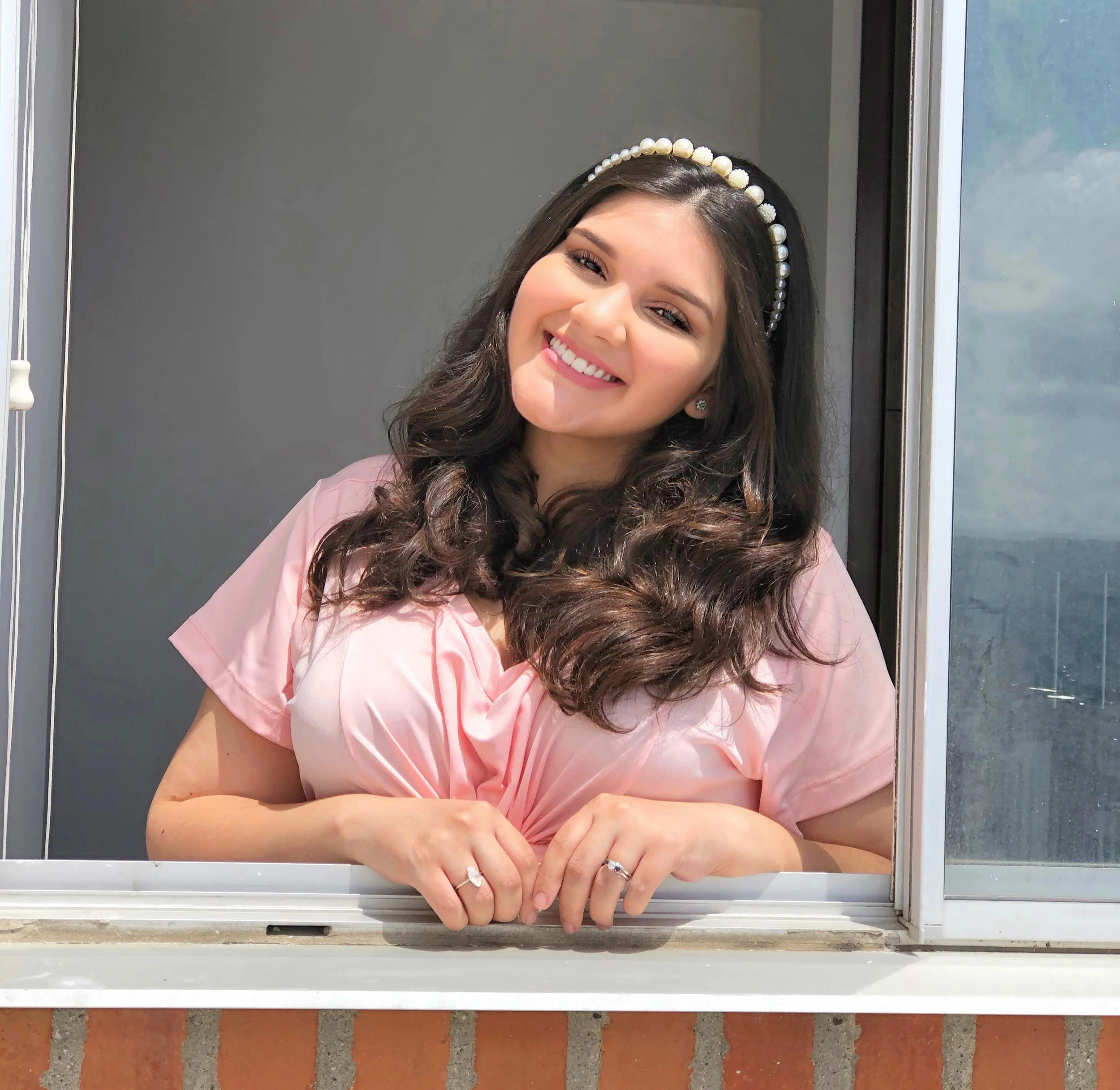 A pregnant woman with long, dark, curly hair leaning out of a window, smiling and wearing a pink top and a pearl headband.