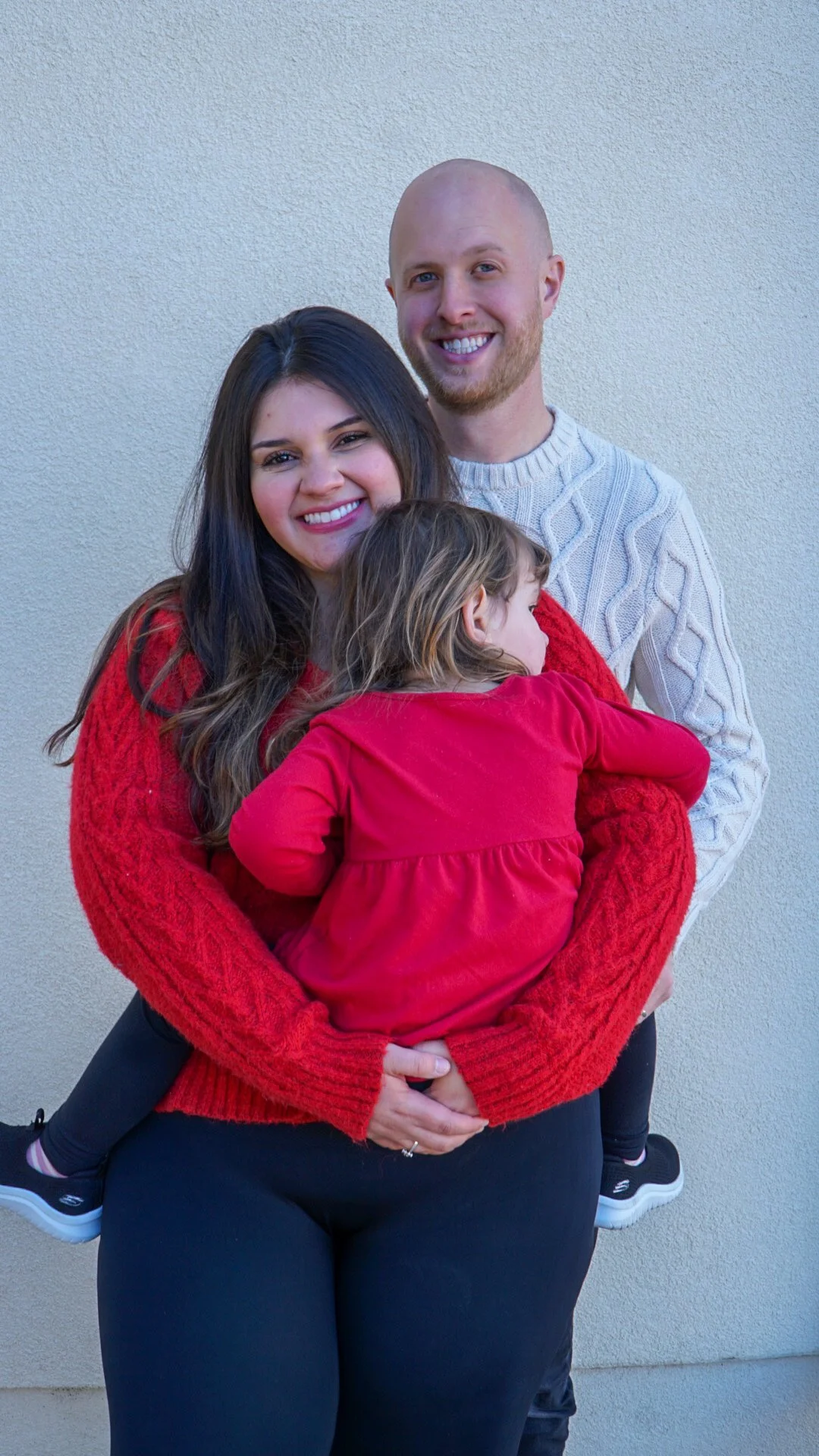 A mother, smiling woman with long dark hair holding a young girl in a red dress, standing next to a smiling bald man in a light gray sweater against a plain light-colored wall.