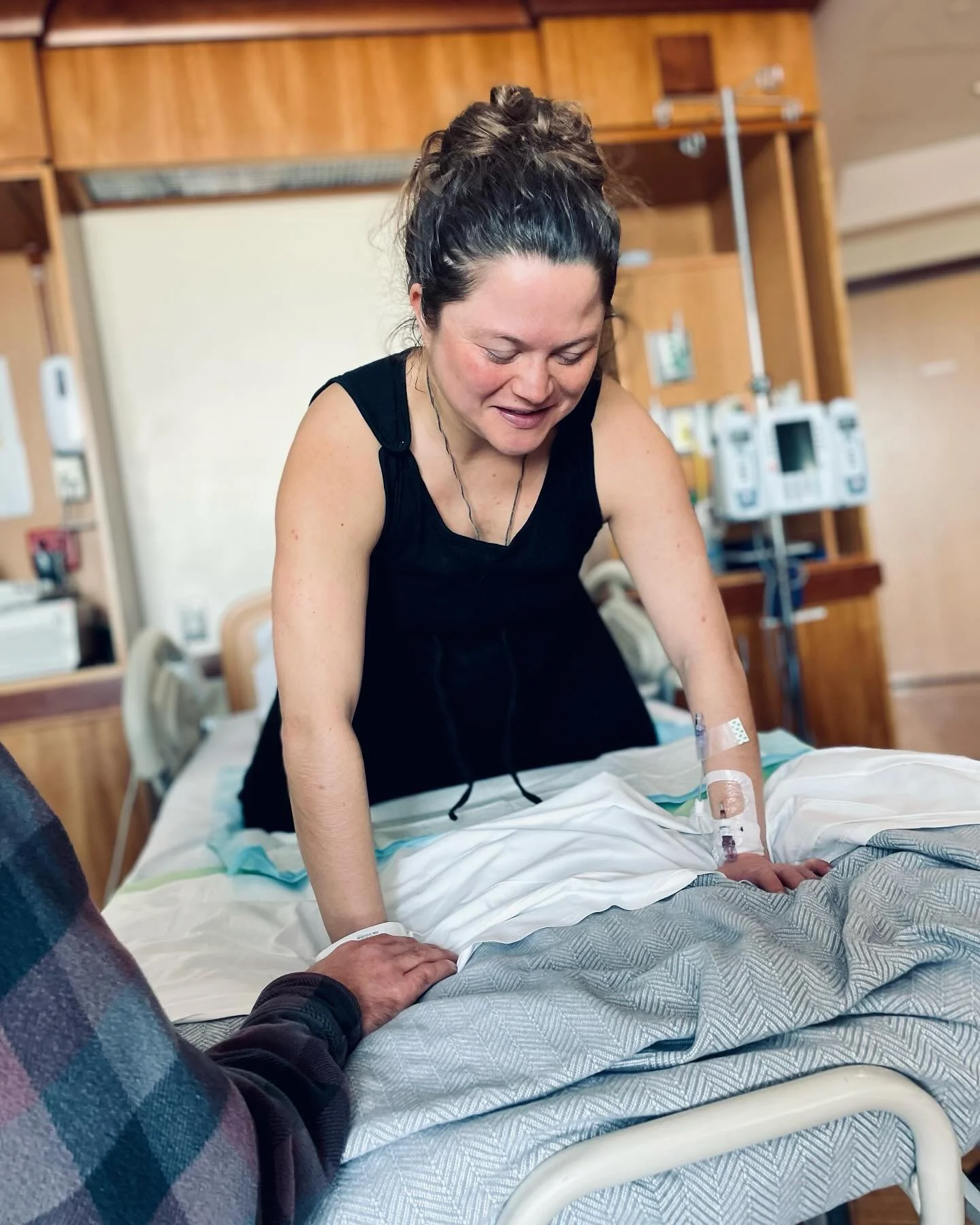 A woman in a hospital room leaning over a hospital bed, trying different labor positions, smiling, with medical equipment visible in the background.