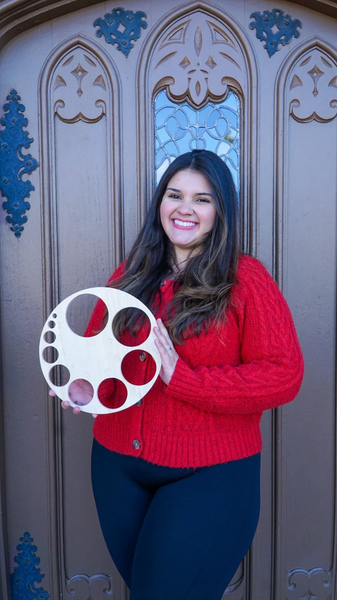 A woman wearing a red sweater standing in front of a decorative wooden door, holding a wooden color palette with circular cutouts that display cervical dilation, smiling at the camera.