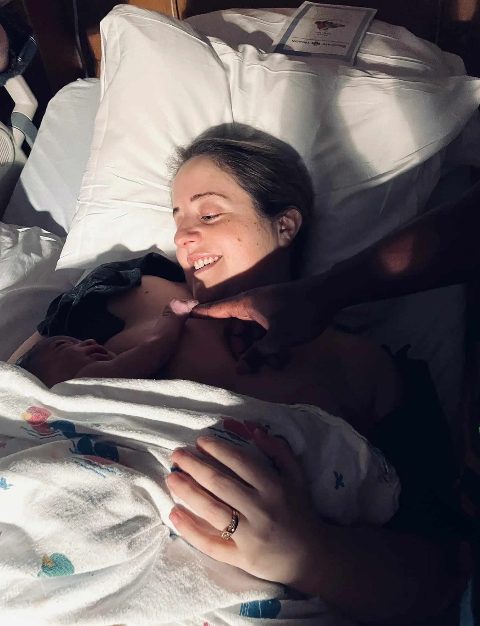 A woman lying in bed, smiling, with her hand gently touching a newborn baby's foot, in a hospital room with white bedding and pillows.
