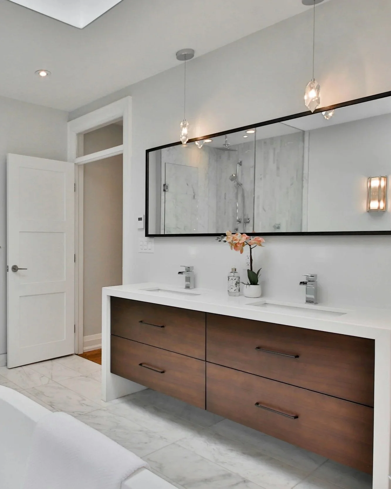 Modern bathroom with dual sinks, large mirror, wooden vanity, marble floors, flowers on countertop, shower in background, minimal lighting.