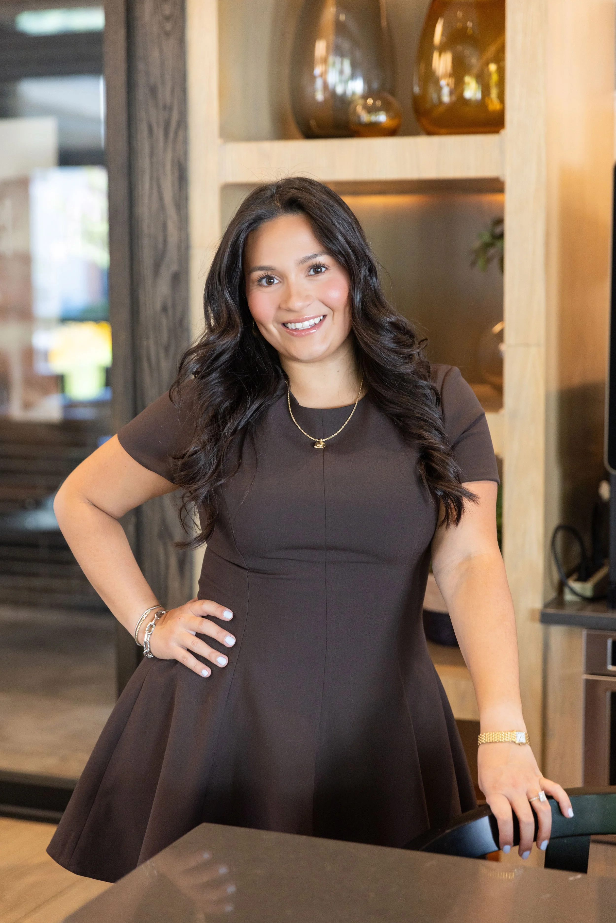 A woman with long dark hair, wearing a dark brown dress, standing in a modern, warmly lit interior, smiling with her hand on her hip.