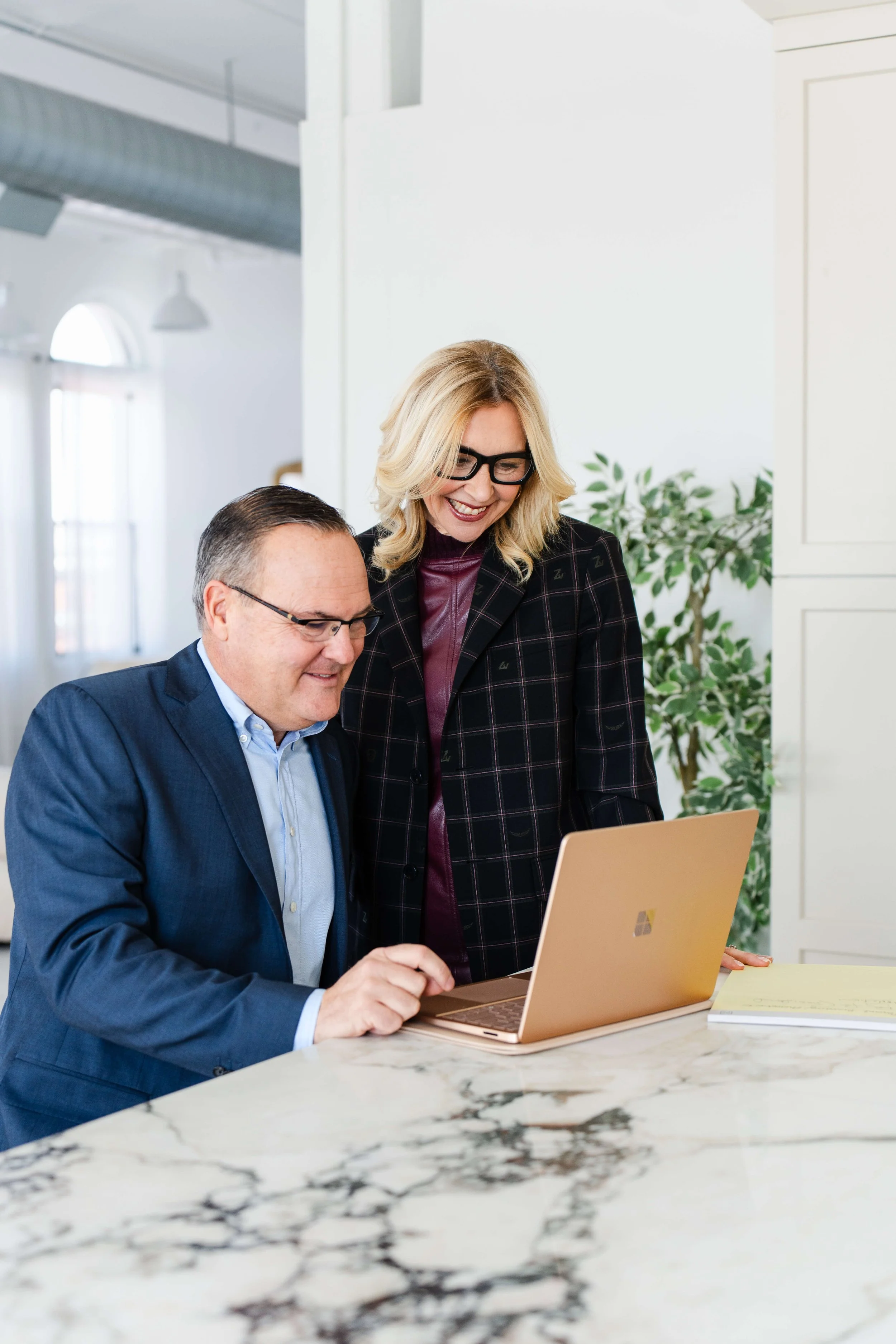 A man in a blue suit and glasses sitting at a marble table using a gold laptop, with a woman with blonde hair, glasses, and a black blazer standing beside him, smiling and looking at the laptop.