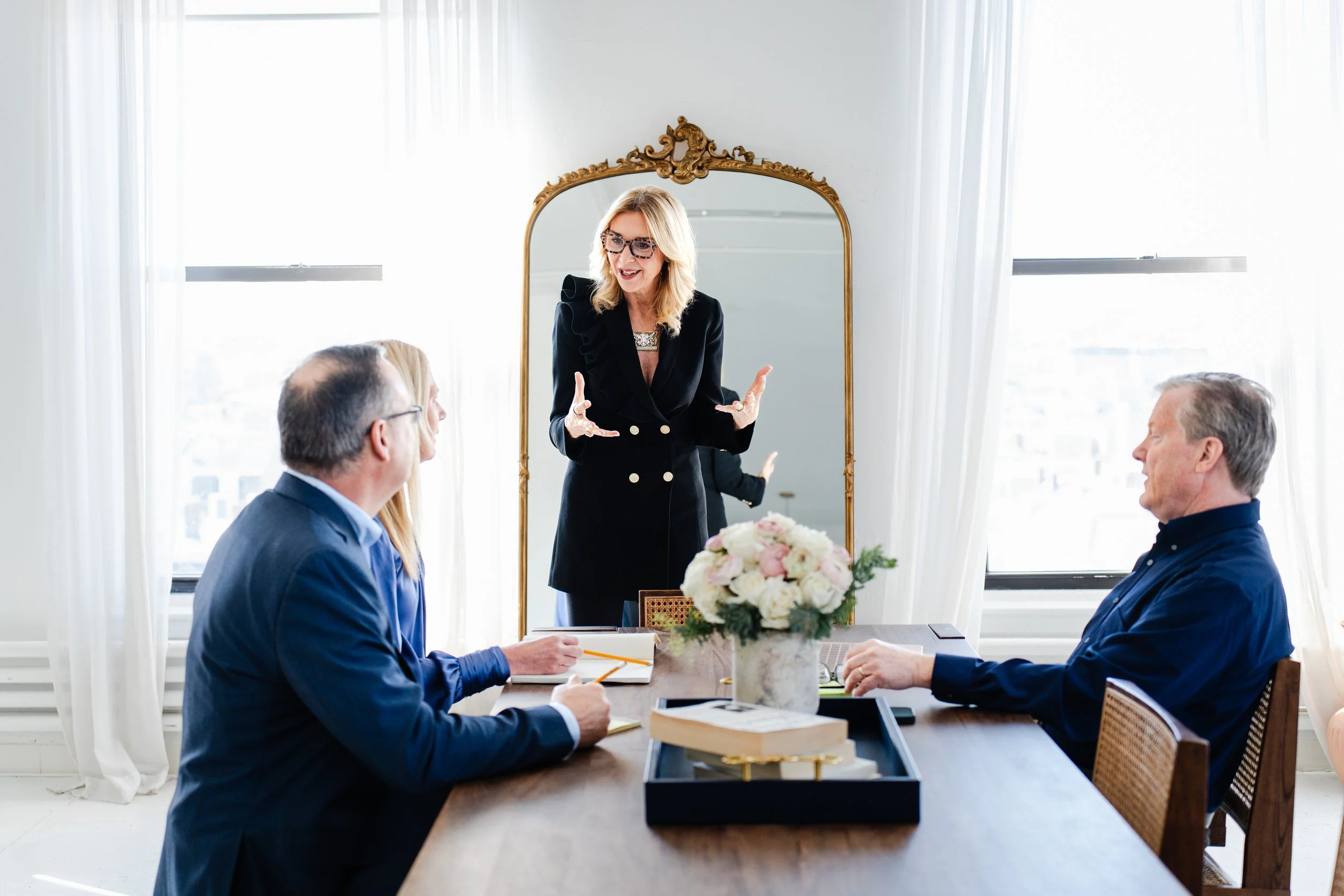 A woman standing and speaking animatedly to four seated people in a bright room with large windows and white curtains, with a gold-framed mirror reflecting her in the background.