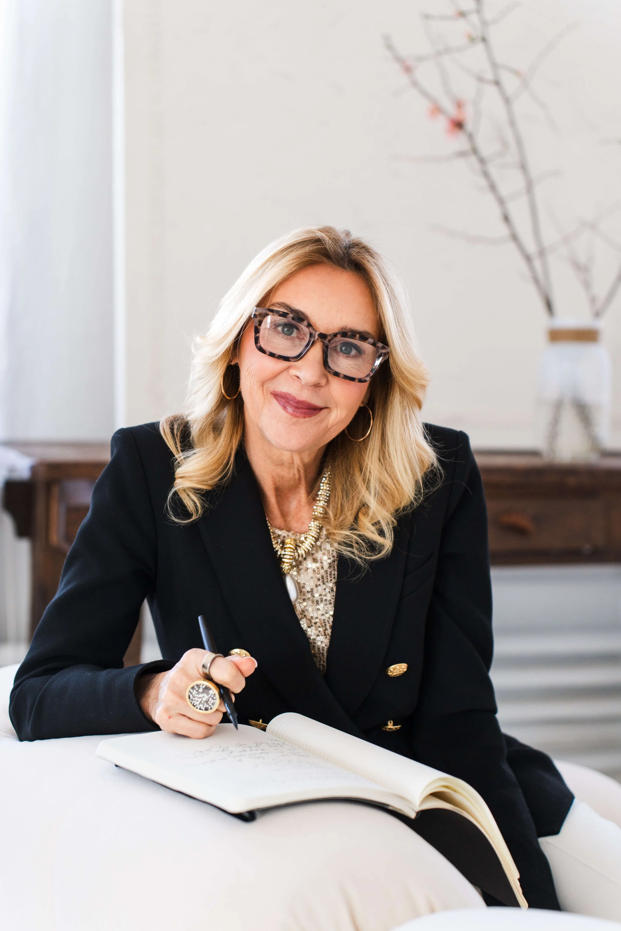 A woman with blonde hair, wearing large tortoise shell glasses, a black blazer with gold buttons, and a pearl necklace, sitting at a white desk with an open notebook, holding a pen, and smiling at the camera.