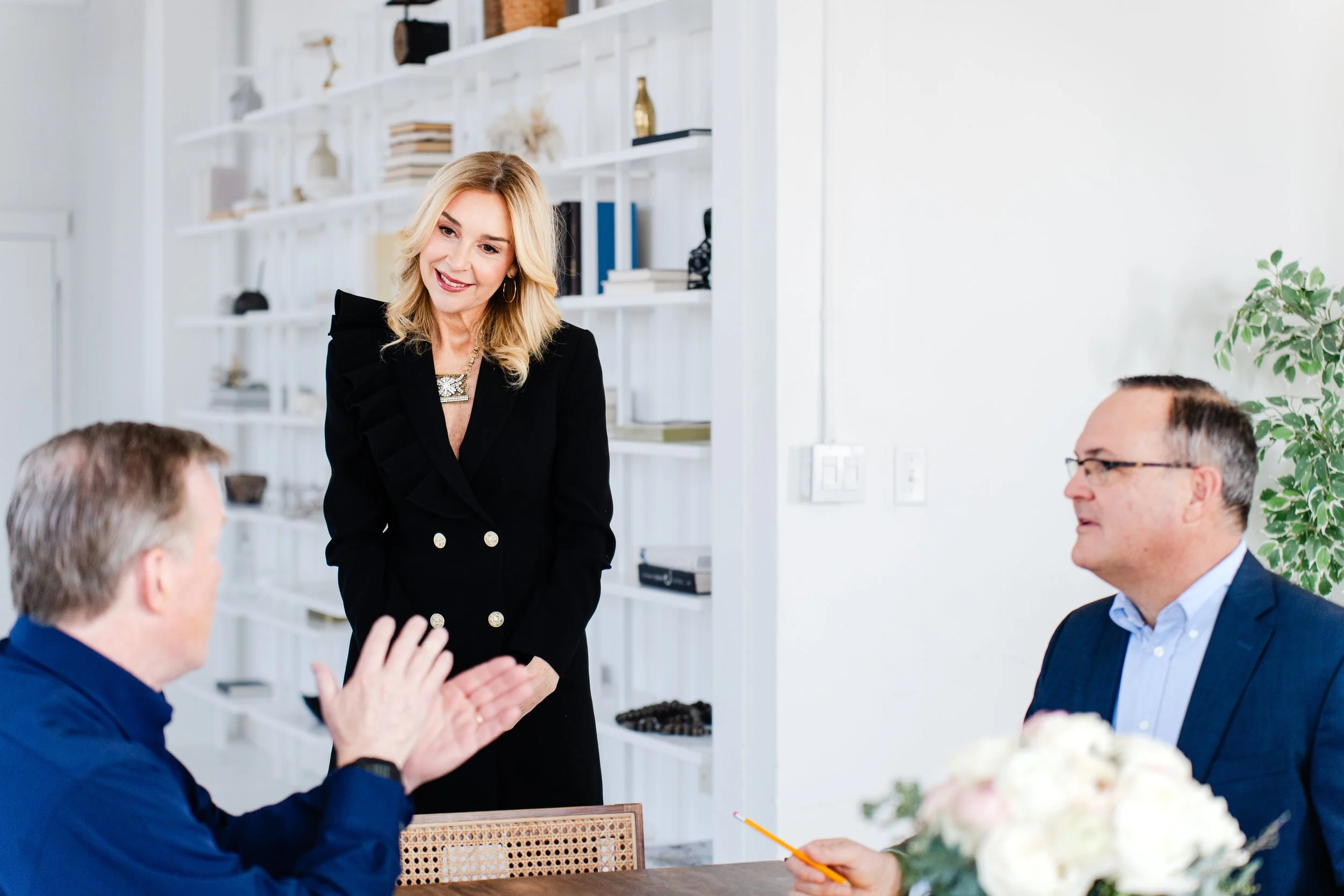 A woman with blonde hair wearing a black blazer stands and interacts with three seated men during a meeting or celebration in a modern, well-lit room decorated with white shelves and a green plant.