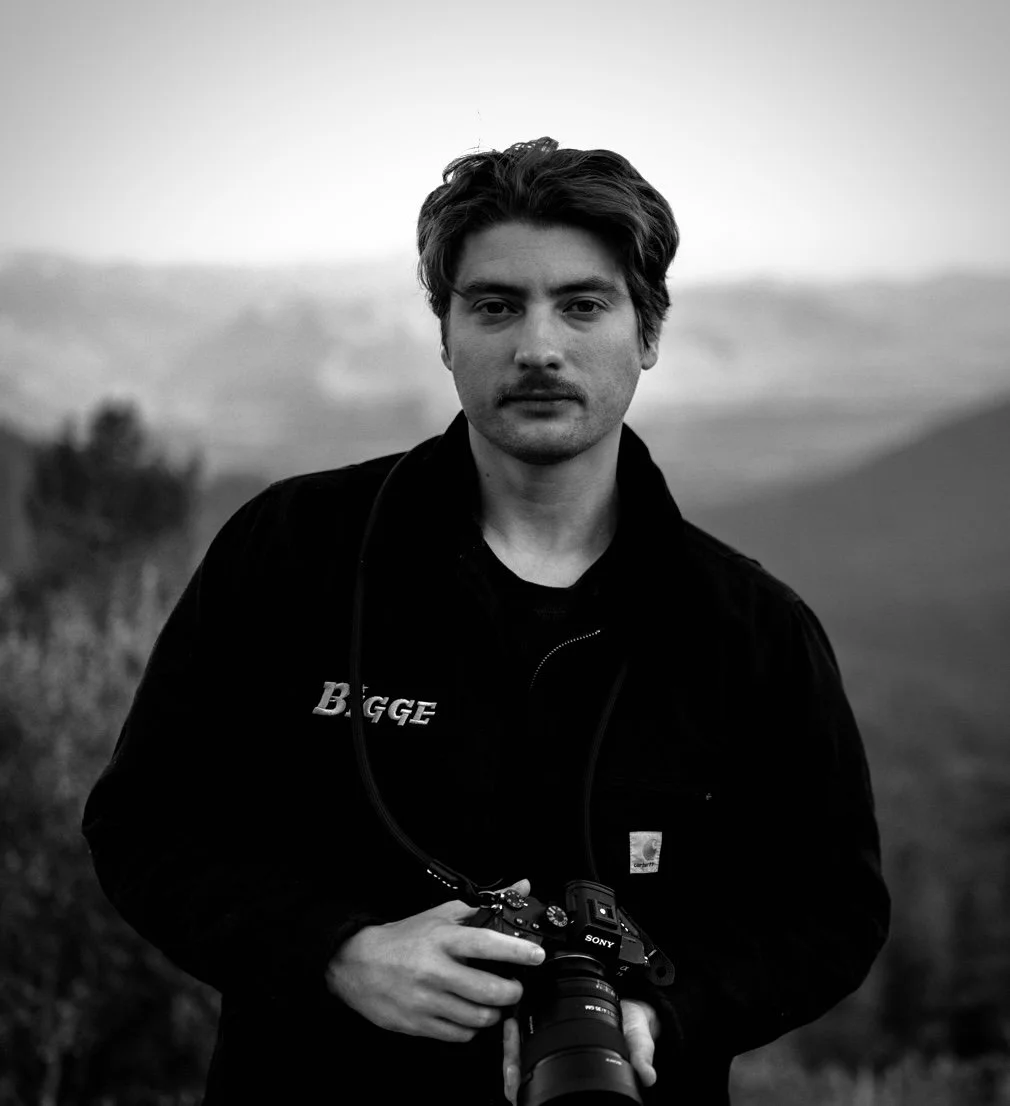 A young man in a dark jacket holding a camera in an outdoor setting with a blurred landscape background in black and white.