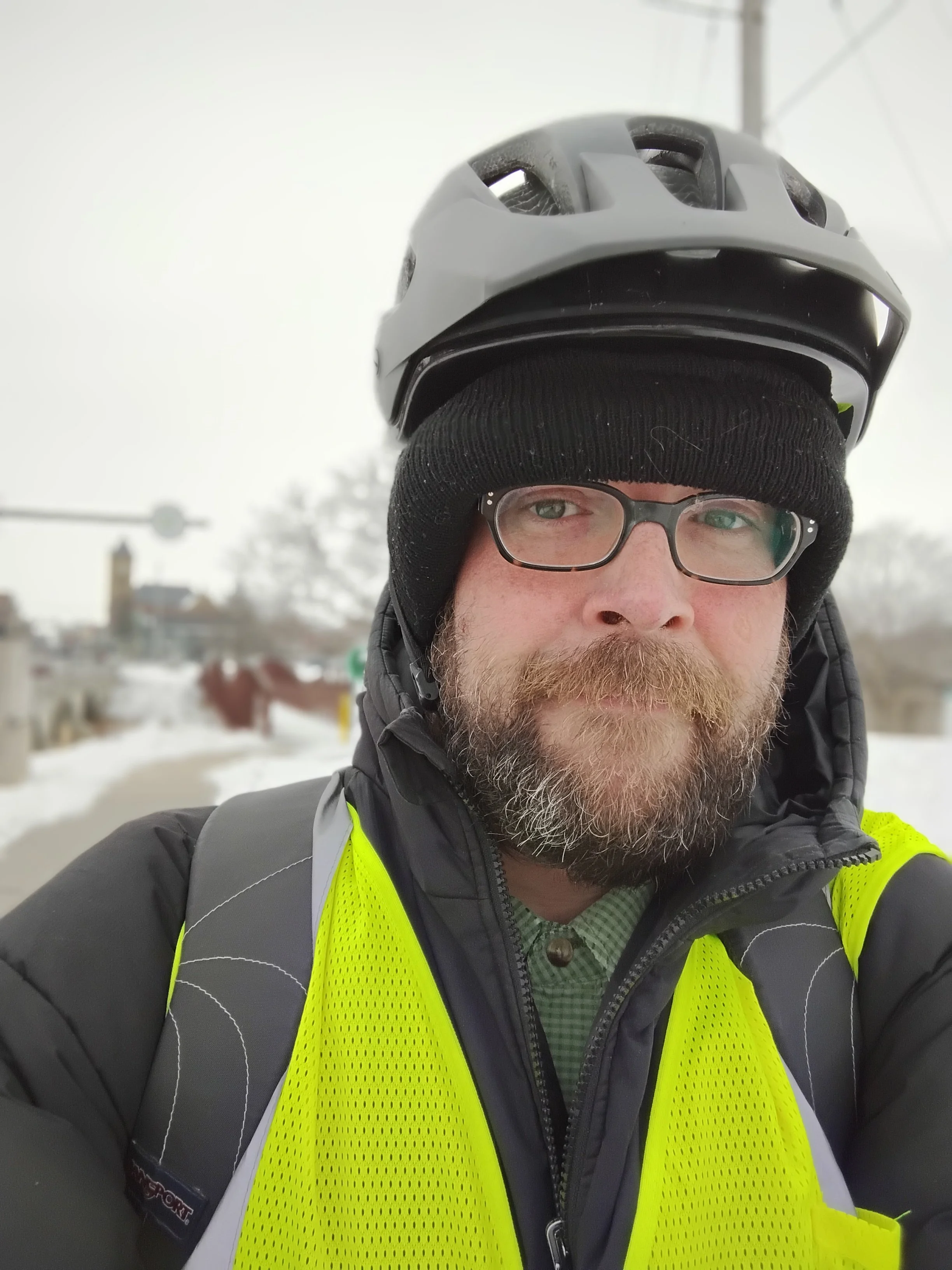 Close-up of a man wearing a bicycle helmet, black beanie, glasses, and a yellow safety vest in a snowy outdoor setting.