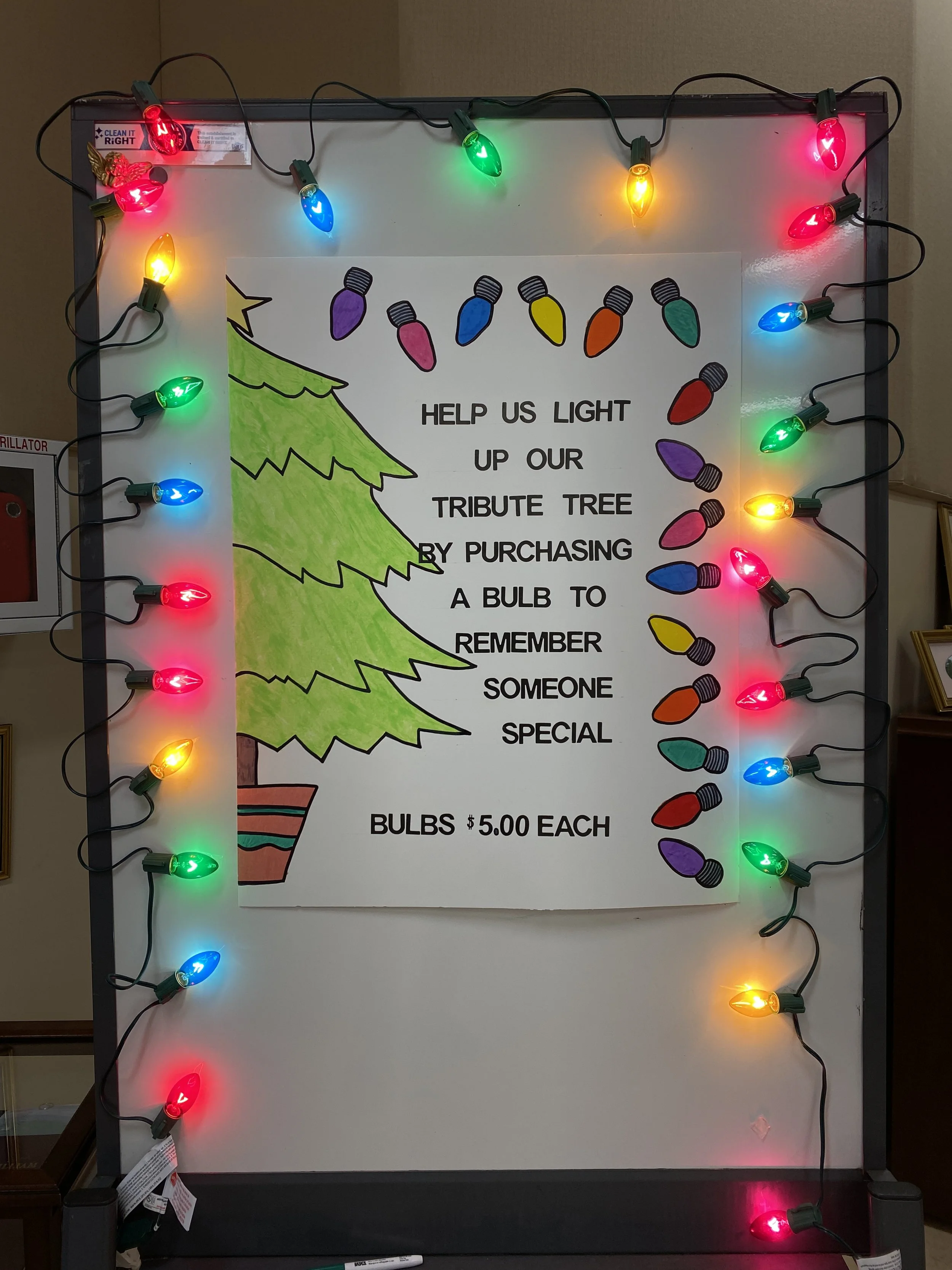 A fund raising poster is framed by a string of Christmas lights that will adorn our outdoor Tribute tree in celebration of loved ones.
