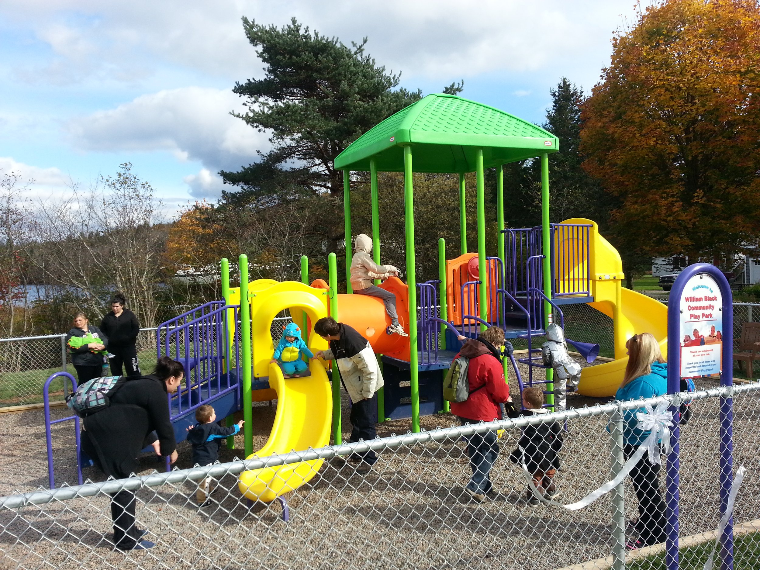 Children and adults playing on a colorful playground at Williams Black Community Play Park, surrounded by a chain-link fence on a partly cloudy day with trees showing fall foliage.