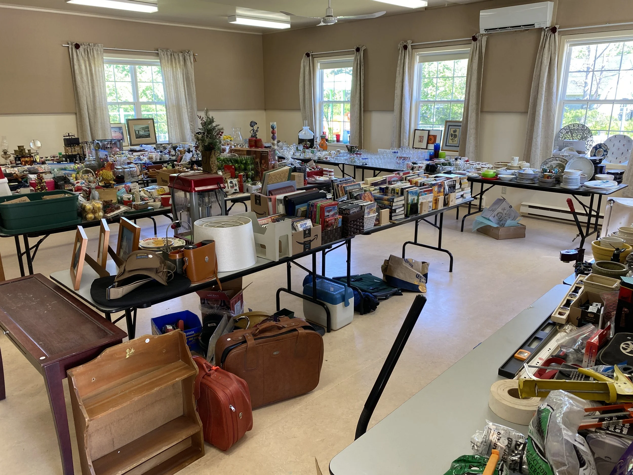 Tables filled with various household items, books, glassware, dishes, and miscellaneous objects inside a brightly lit room, likely set up for a sale or donation event.