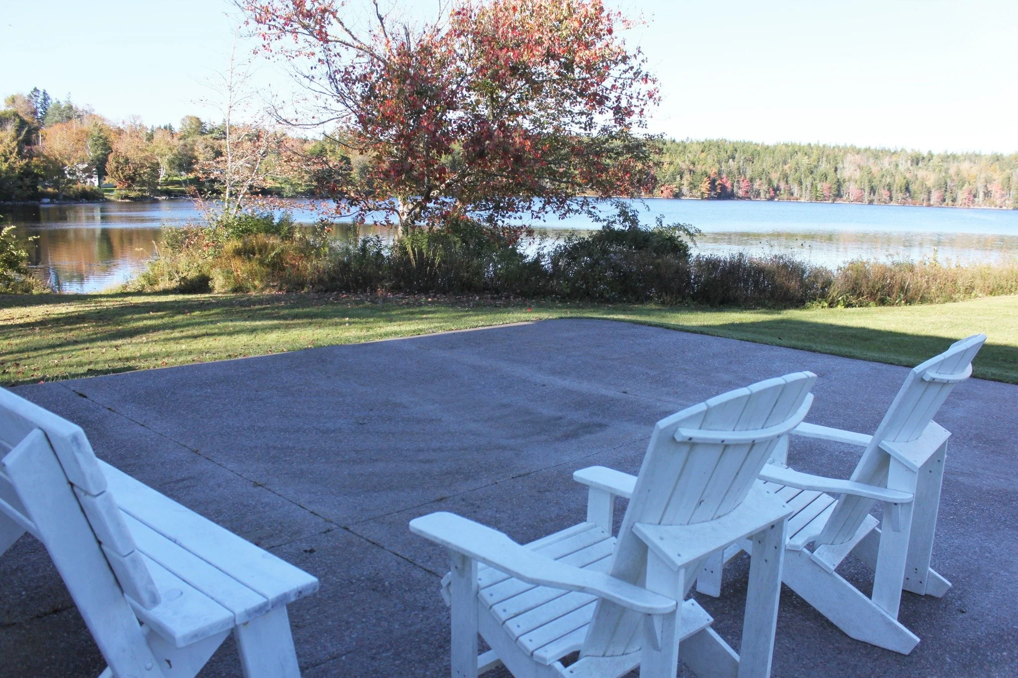 Two white Adirondack chairs and a white bench on a concrete patio overlooking a lake with trees in the background.