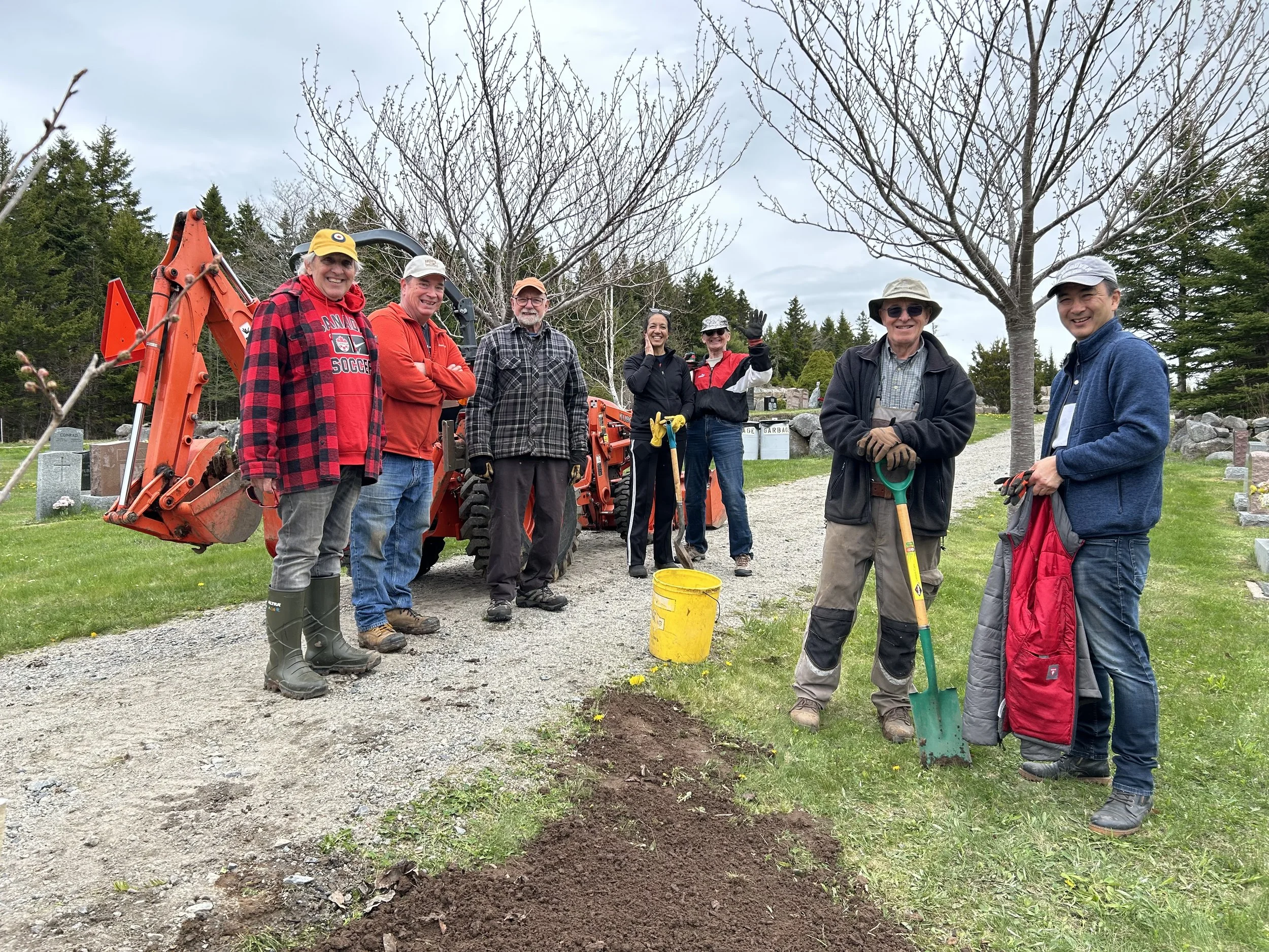Group of eight people planting a tree in a cemetery with gravestones in the background on a cloudy day.