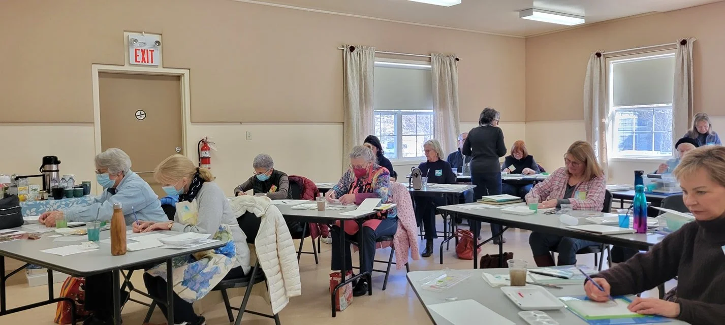 A classroom with adults sitting at tables writing or working on papers, many wearing face masks. There are windows with curtains, and a fire extinguisher near the door. The room has bright natural light.