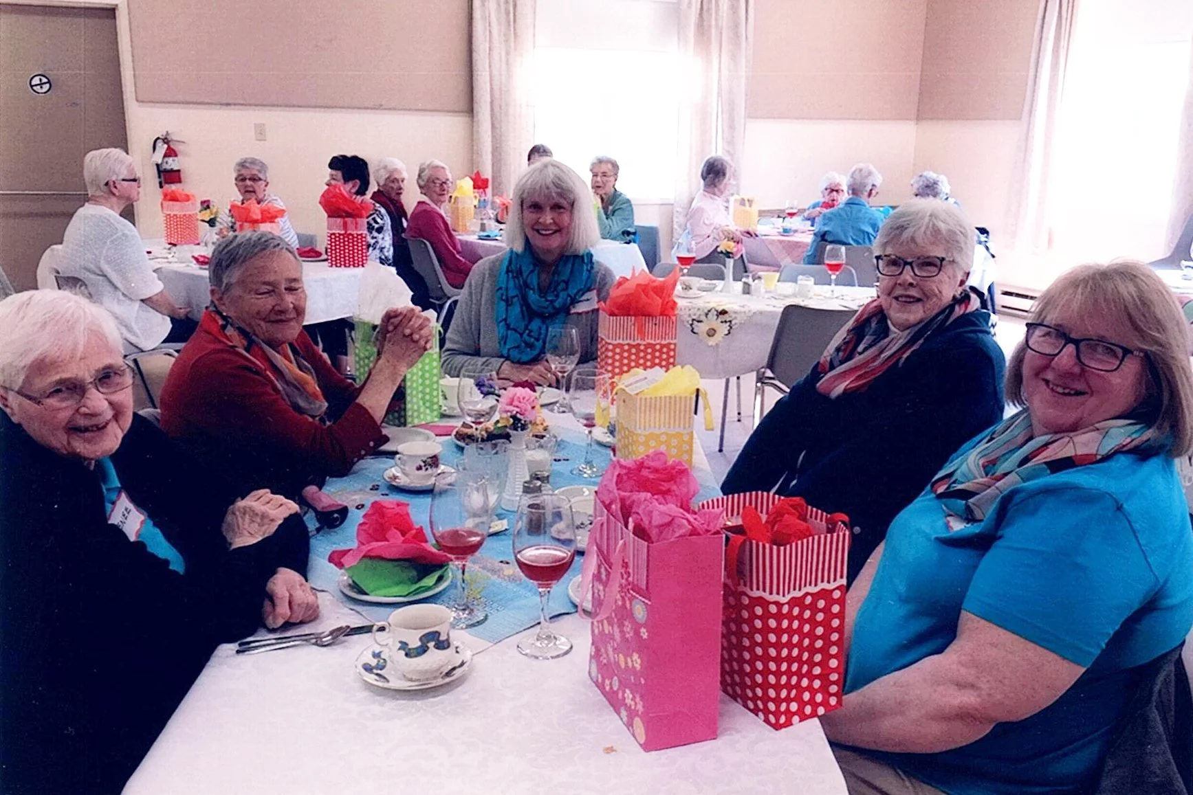 A group of elderly women celebrating at a party in a dining hall, sitting around a decorated table with gift bags, flowers, and drinks.