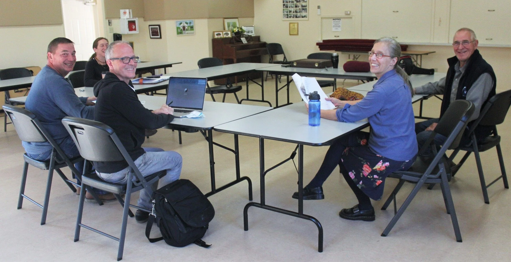 A group of six people sitting around a rectangular table in a meeting room, smiling and looking at the camera. Four people are on the left side, two on the right. The room has chairs, a piano, and framed artwork on the walls.