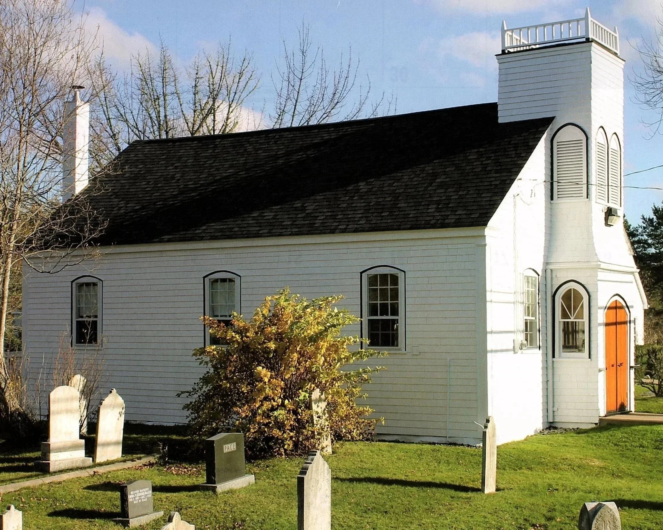 A side view of William Black Memorial United Church glowing white in the sunshine, surrounded by old gravestones casting long shadows on the well tended lawn.