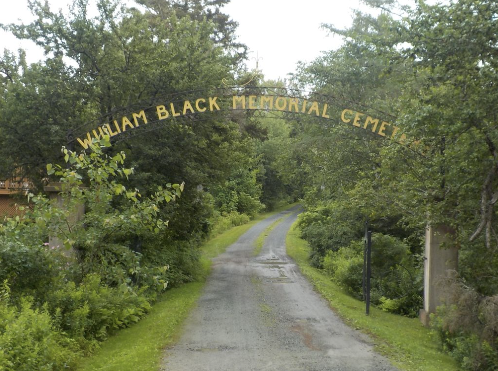 A black wrought iron arch on stone pillars bearing the words William Black Memorial Cemetery spans a gravel drive leading into a cluster of trees. 