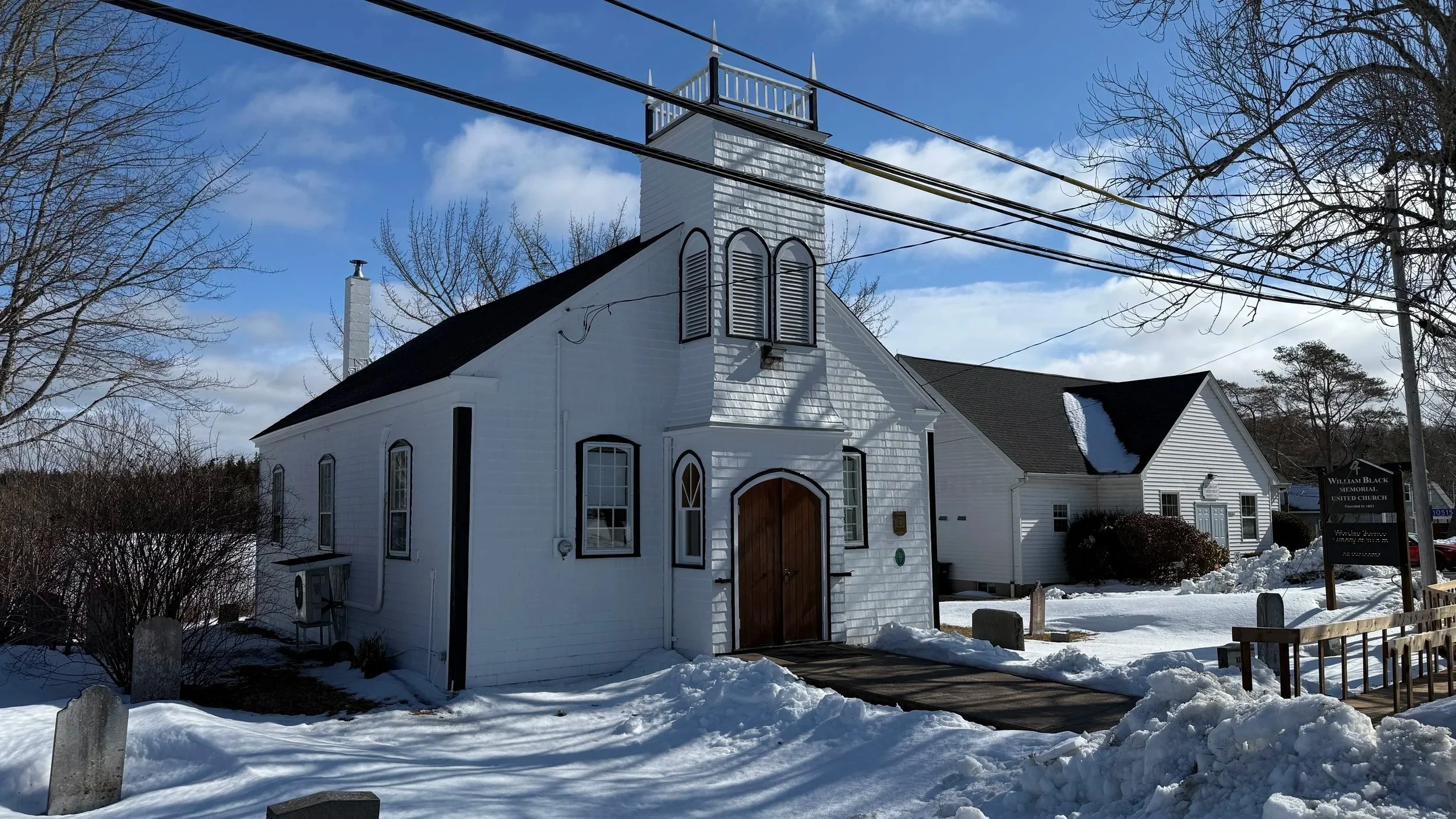 The little white wood shingled church in Glen Margaret with a new electronic sign and church hall in the background. There is a small steeple and the walkways are lined by snowbanks on a sunny winter day.