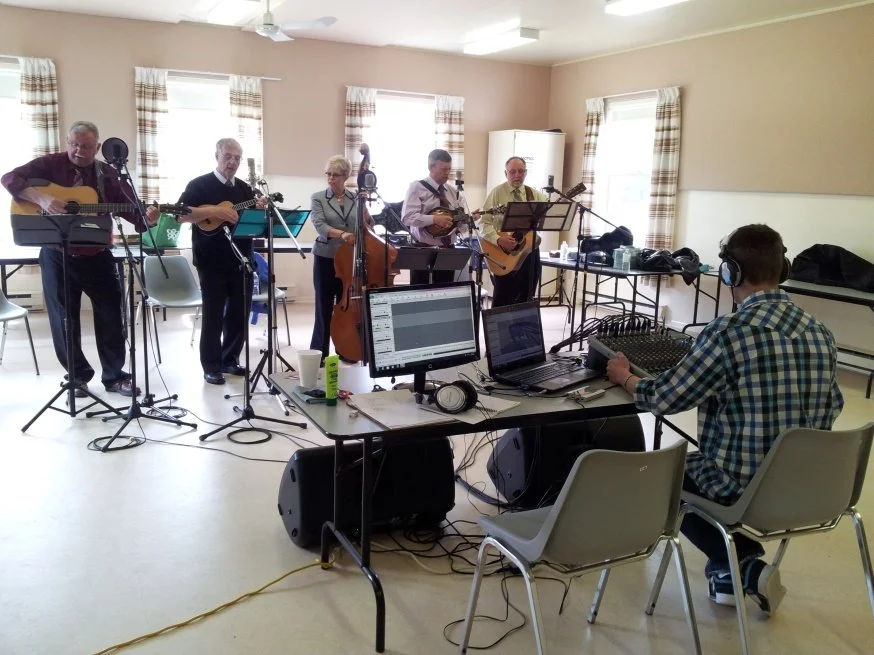 A group of six musicians playing guitars and a double bass in a room with windows, while a sound technician uses a laptop and audio equipment to record or mix the music.