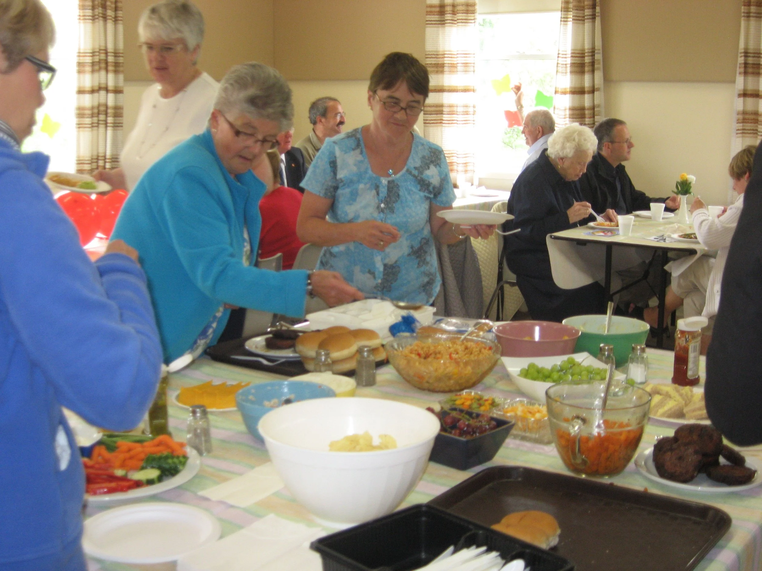 People serving and preparing food at a buffet-style gathering in a decorated room with window curtains and colorful paper cutouts on the windows.