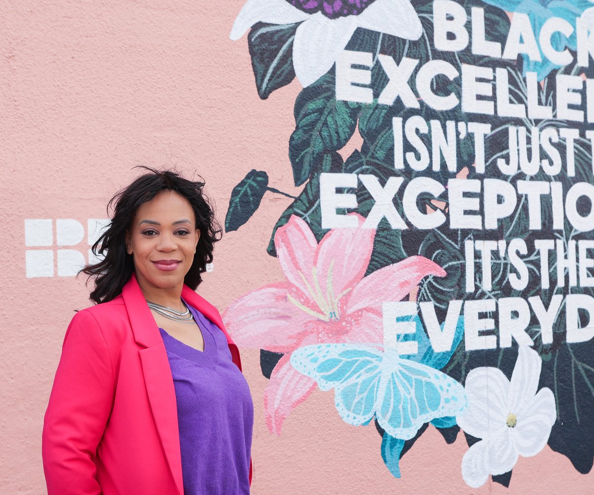 A woman with medium dark skin and black hair standing in front of a pink wall with painted flowers and a quote, wearing a pink blazer over a purple top.