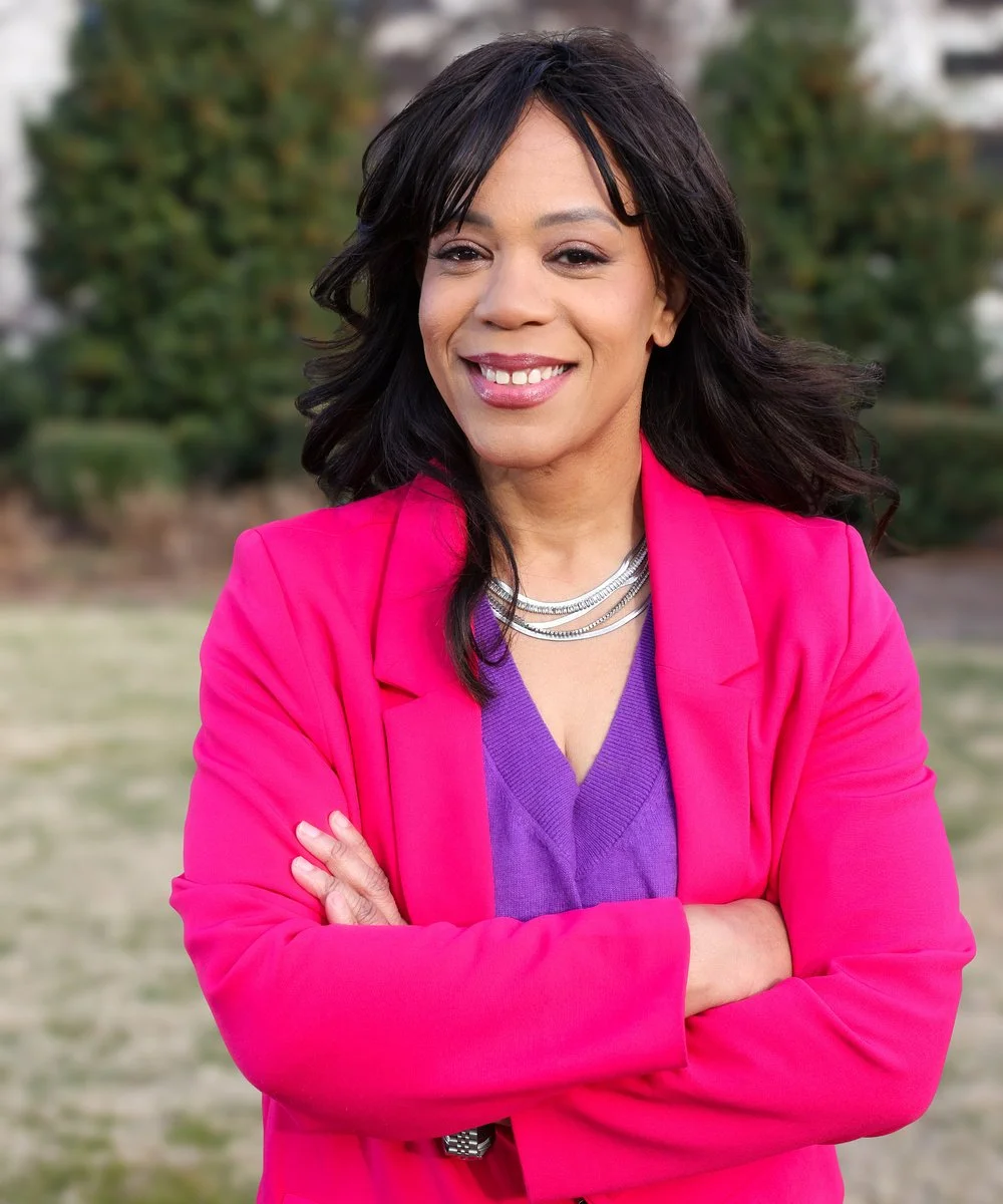 A woman smiling outdoors with arms crossed, wearing a pink blazer over a purple top, and silver necklaces. Background is blurred with trees and grass.