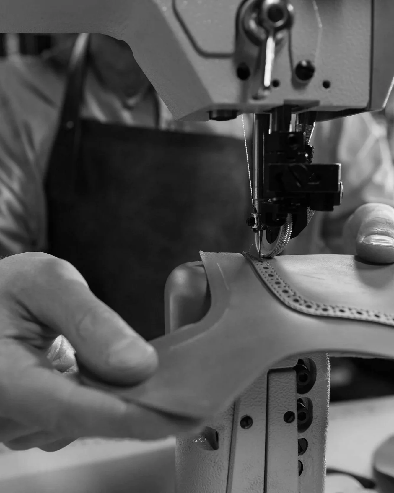 A close-up black and white photo of a person operating a sewing machine, sewing a piece of fabric with decorative perforations.