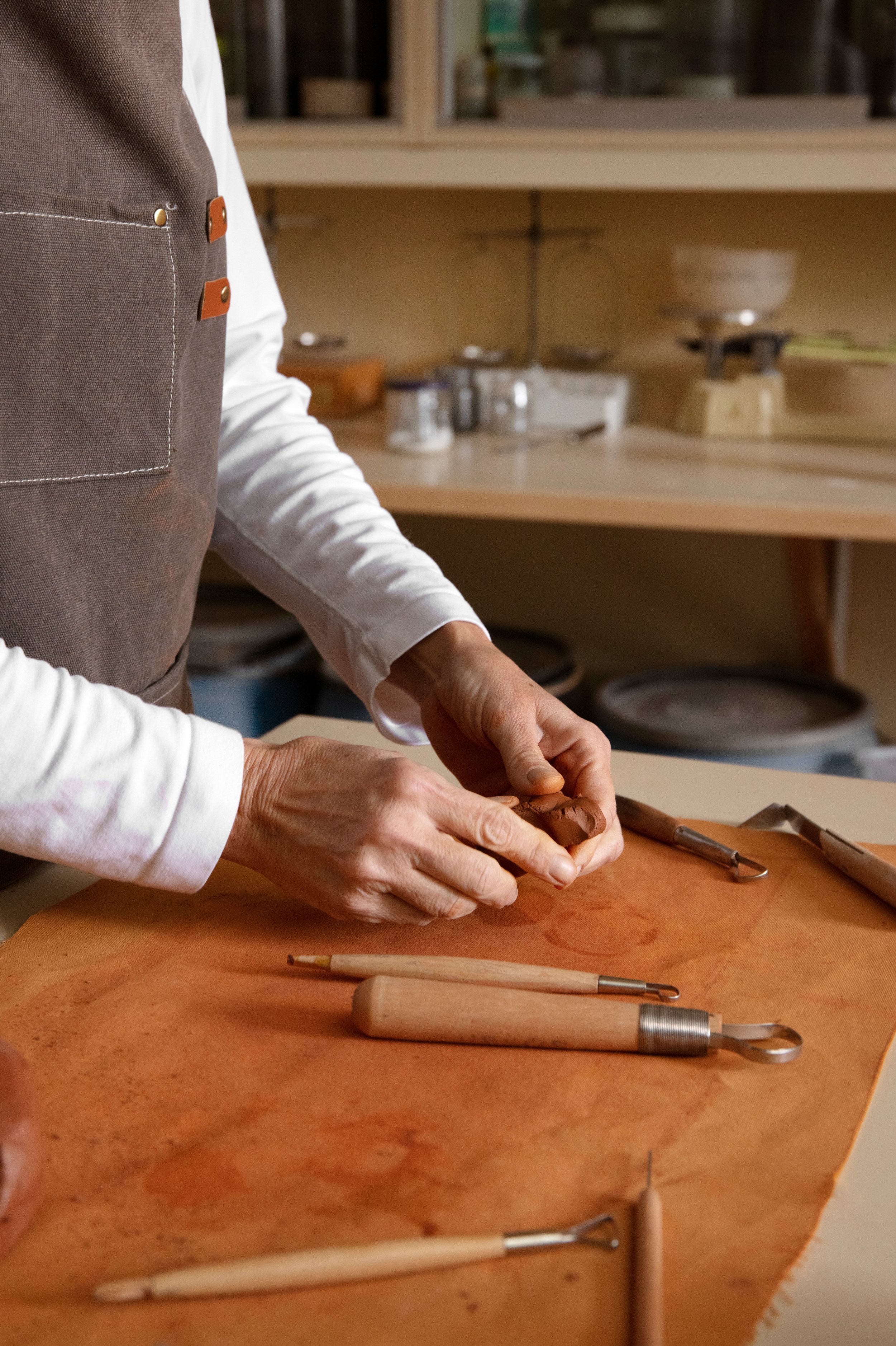 Individual shaping clay on a work surface in a pottery studio, with pottery tools nearby.