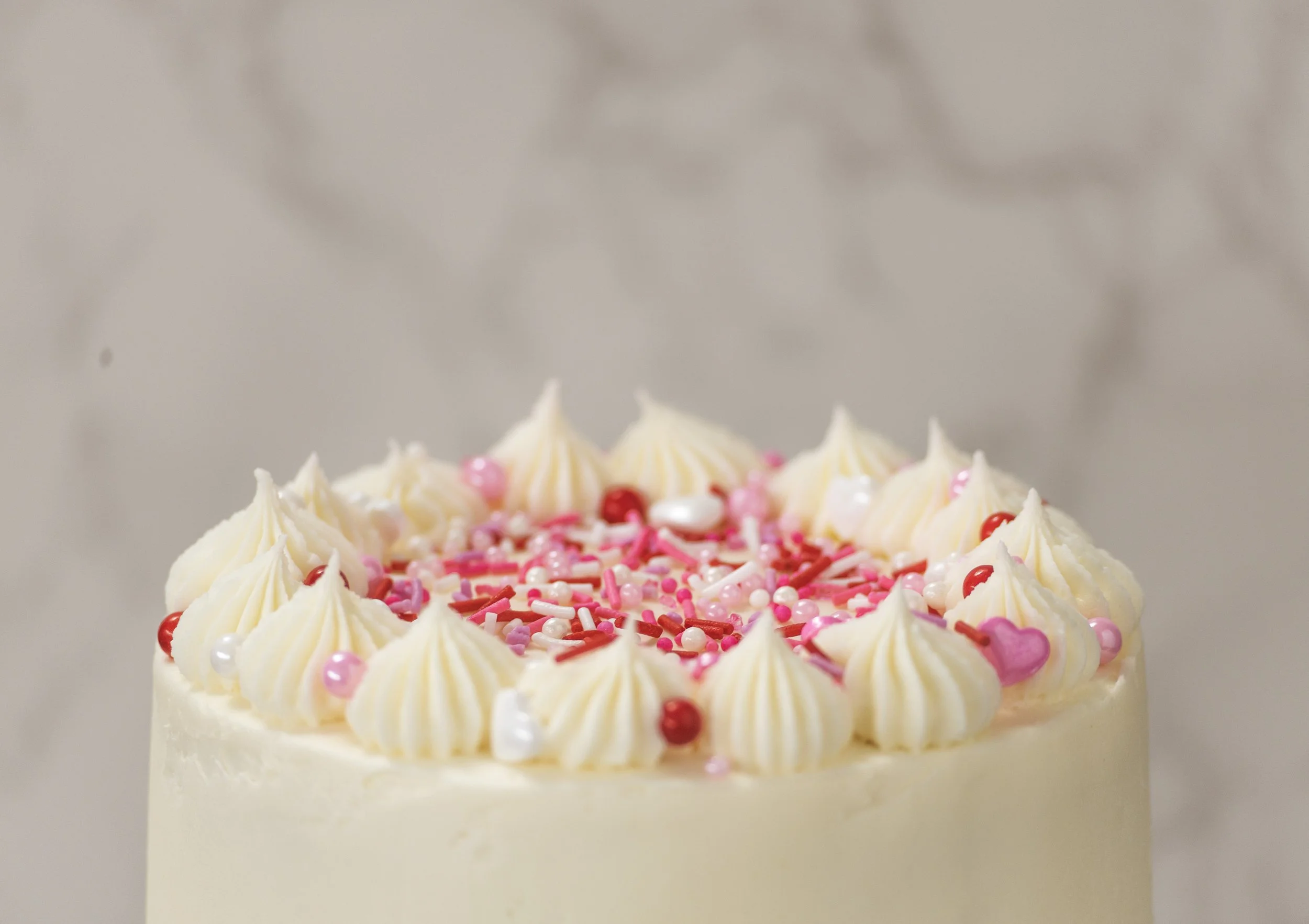 A decorated birthday cake with white frosting, pink and red sprinkles, and pink heart-shaped decorations.