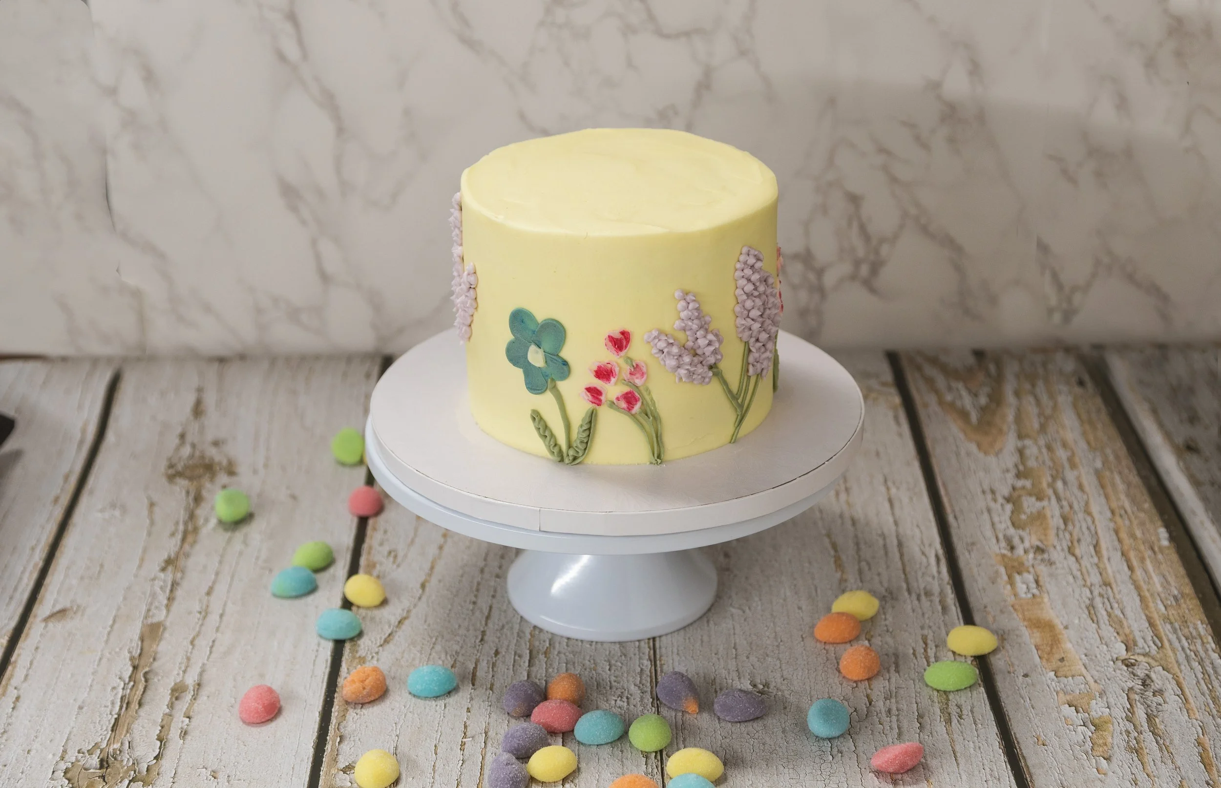 A small yellow cake with floral designs on the side, placed on a white cake stand. Colorful candies are scattered around the cake on a rustic wooden surface.