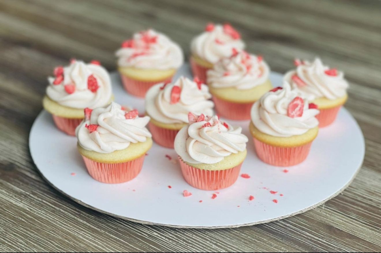A white plate with cupcakes topped with pink and white swirled frosting and red sprinkles, set on a wooden surface.