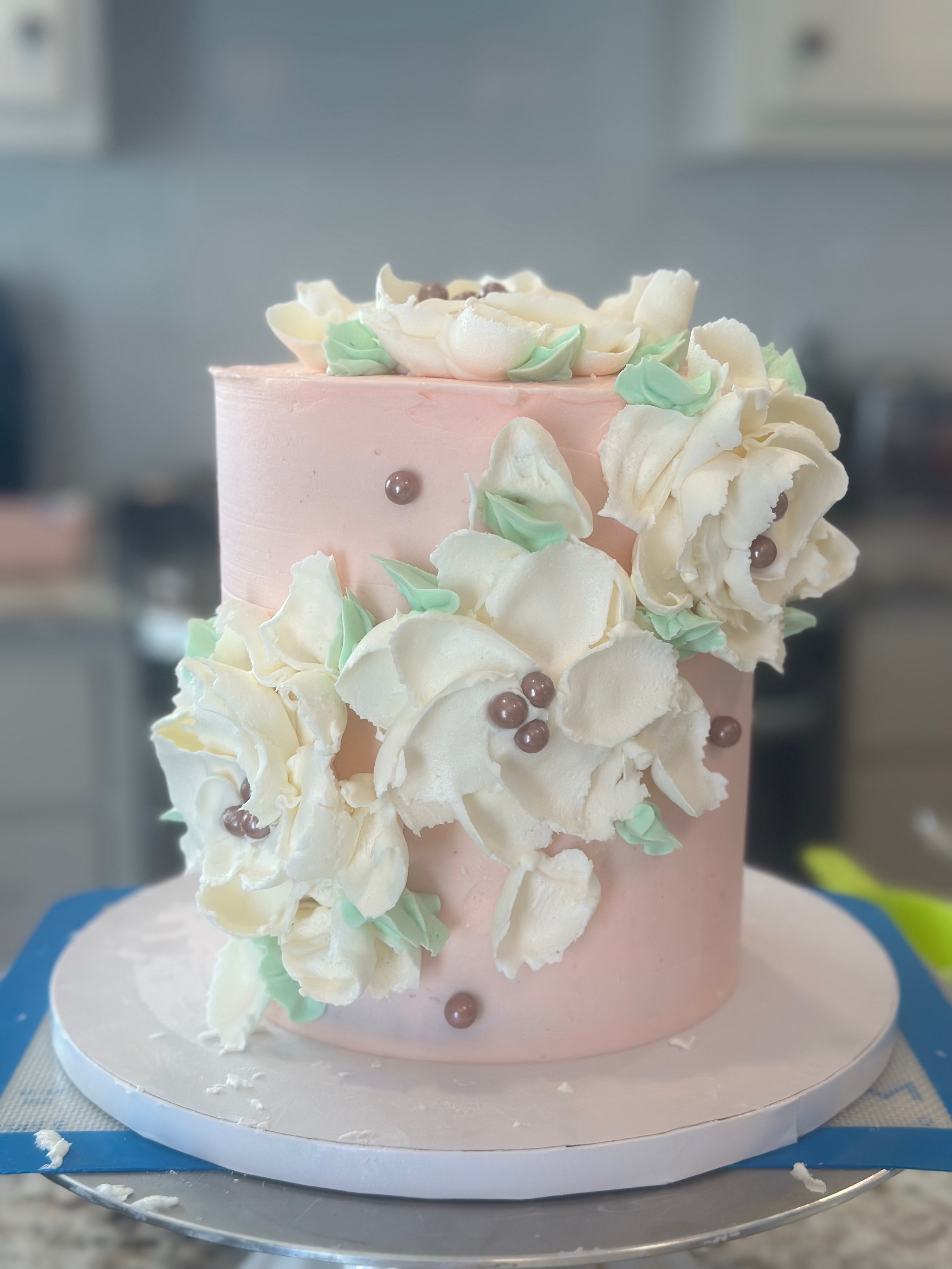 A pink cake decorated with white and green flower-shaped icing, topped with rose gold pearls, on a white cake board.