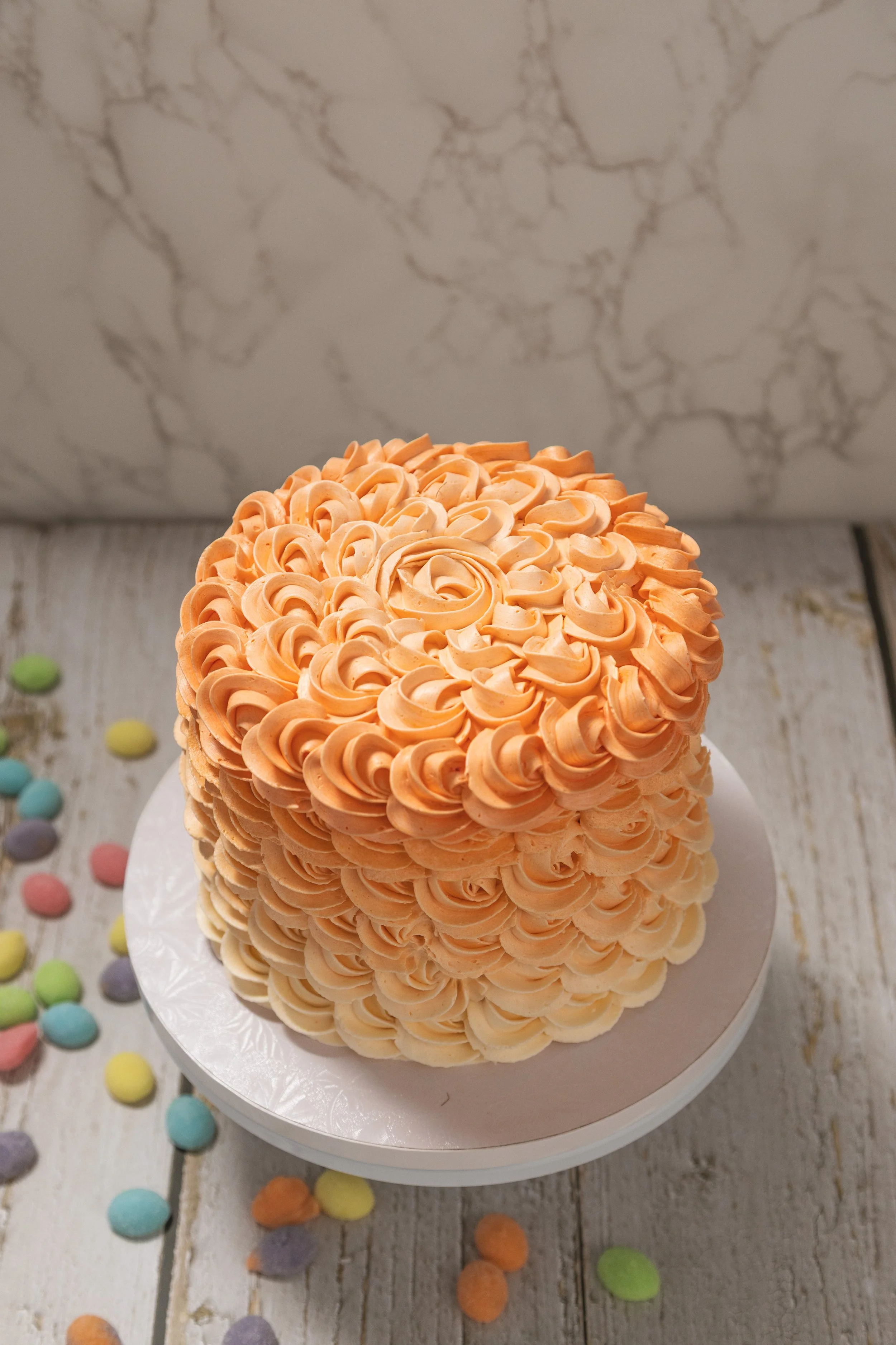 A round cake decorated with orange frosting swirls arranged in a floral pattern, placed on a white cake stand. Colorful candy-coated chocolates are scattered on a wooden surface around the cake.