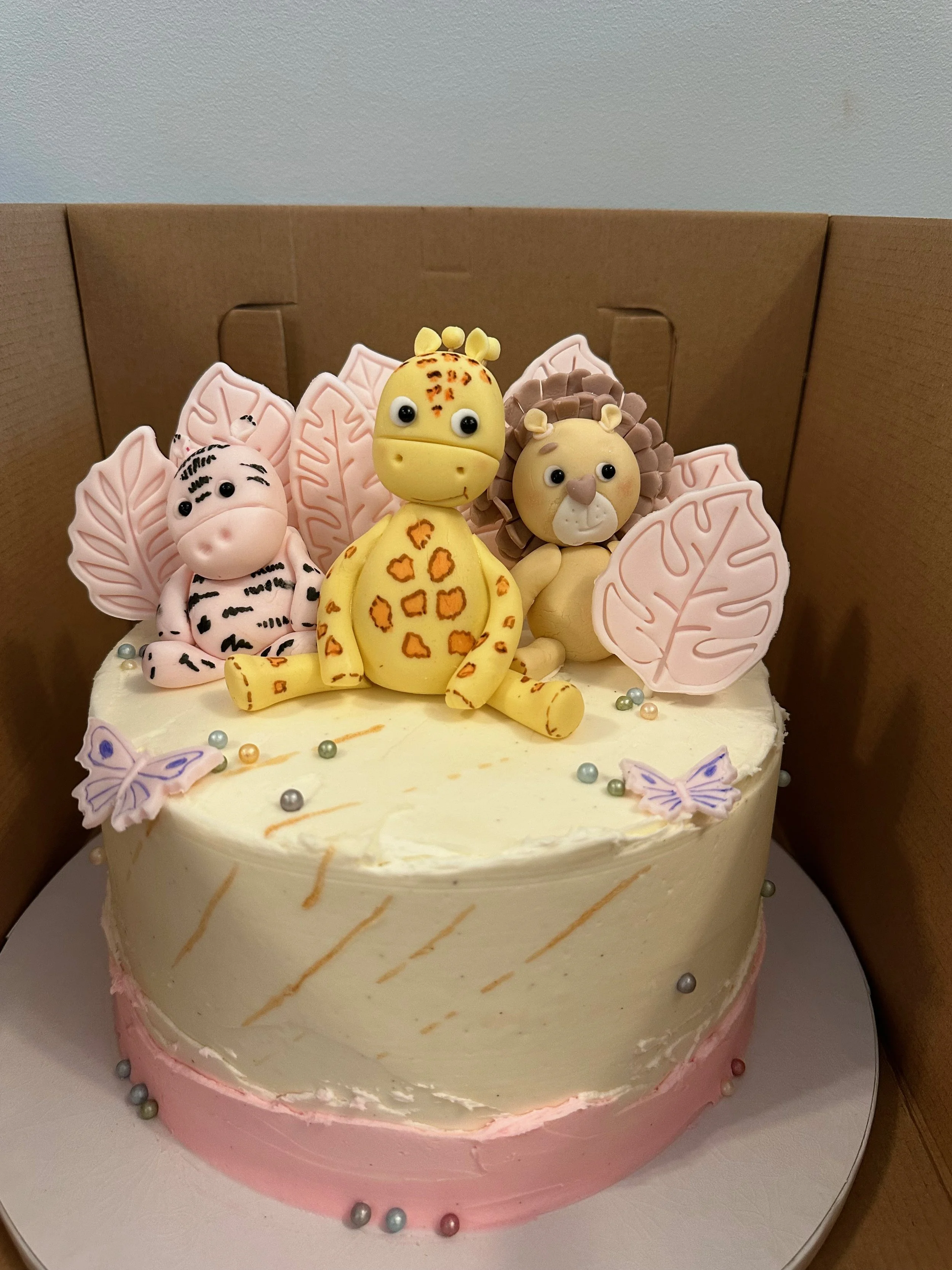 Decorative cake featuring animal and leaf shaped fondant figures, such as a giraffe, lion, and zebra, with pastel pink and white icing, colorful edible pearls, and butterfly decorations.