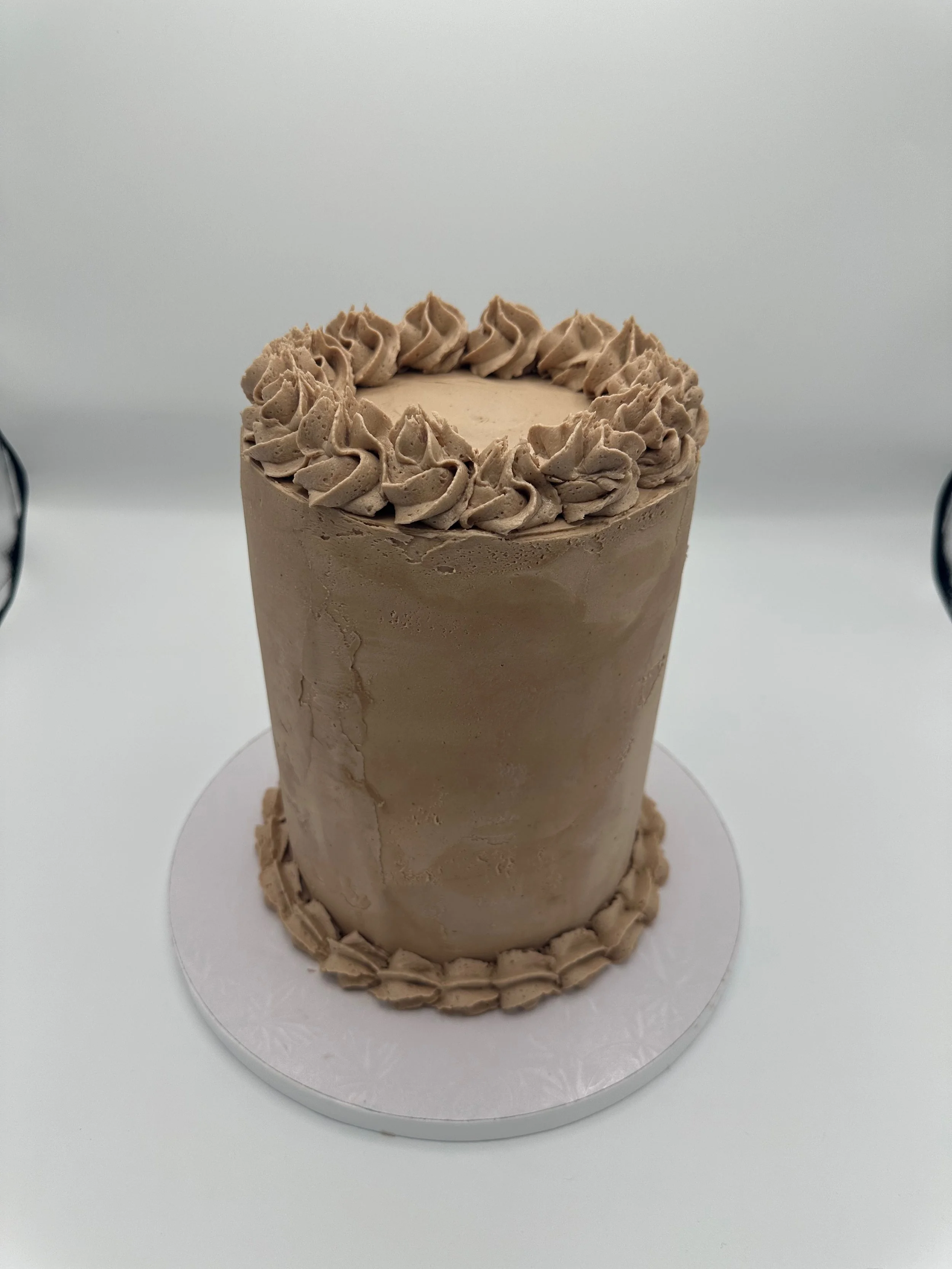 A tall, chocolate cake decorated with piped frosting around the top rim and on the bottom edge, placed on a white surface.