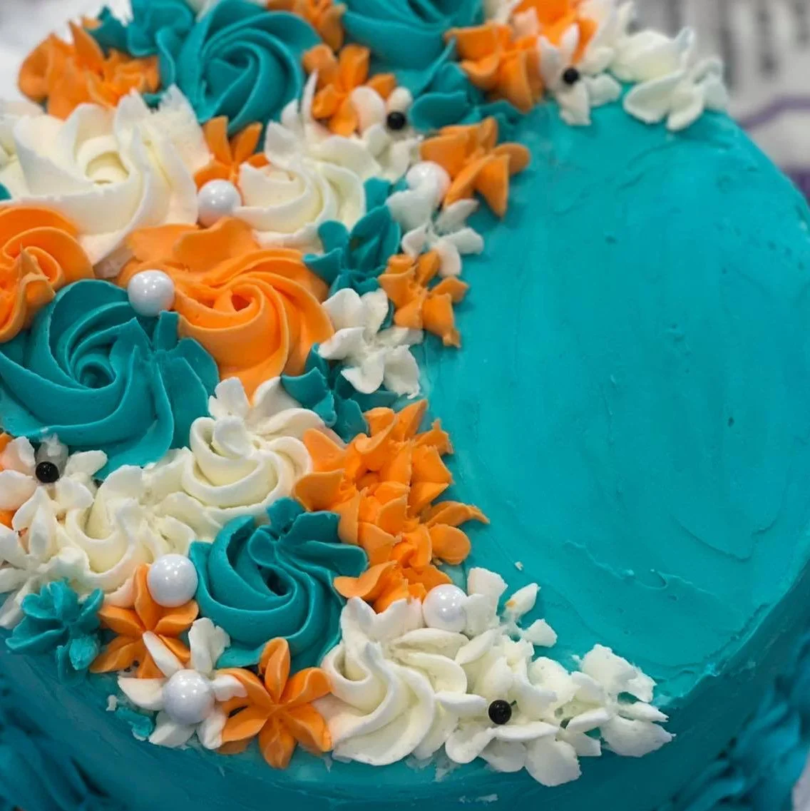 Close-up of a decorative birthday cake with blue, orange, and white frosting flowers, white edible pearls, and small black decorative accents.