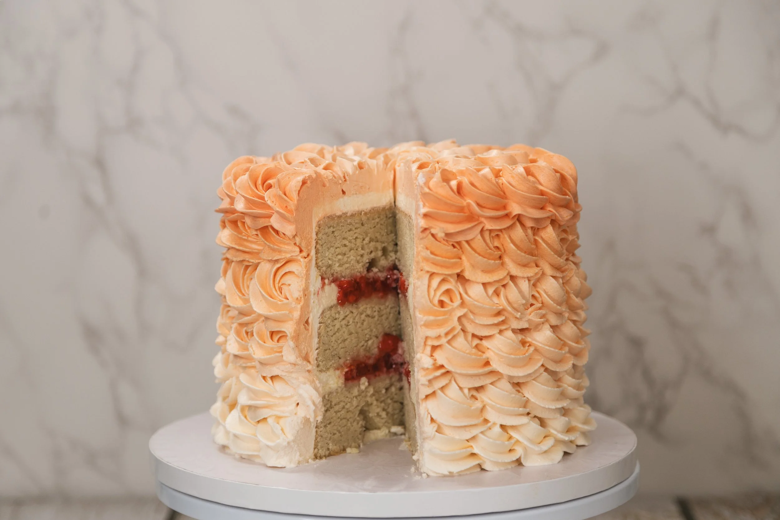 A round cake with orange piped frosting swirls, partially sliced to show layers of light-colored sponge, cream filling, and a red fruit jam at the bottom inside, placed on a white cake stand against a marbled background.
