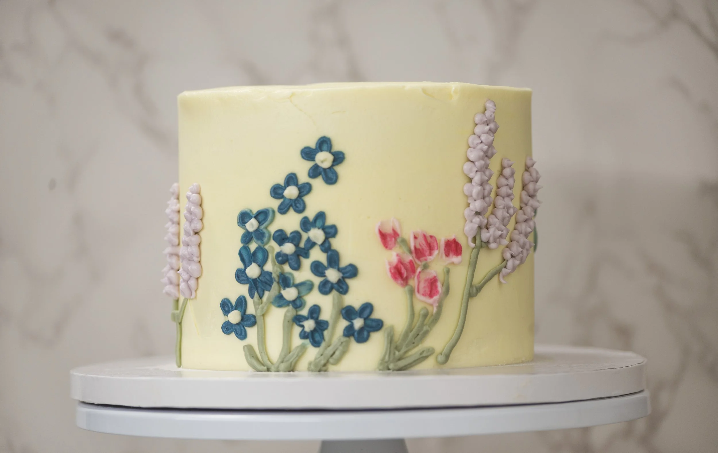 A tall, decorated cake with a yellow icing base. The cake features blue, pink, and purple floral decorations made of icing or fondant, with green leaves and stems. The cake is placed on a white cake stand against a neutral, blurred background.
