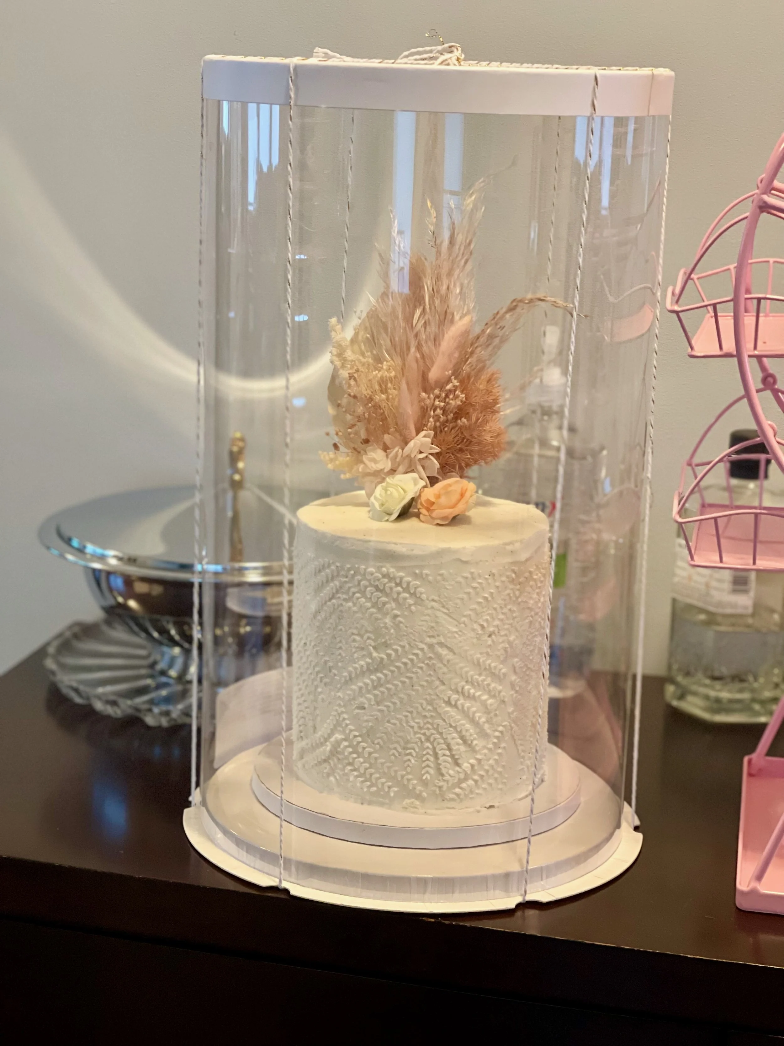 A decorative cake with dried flowers inside a clear plastic case, placed on a dark surface.