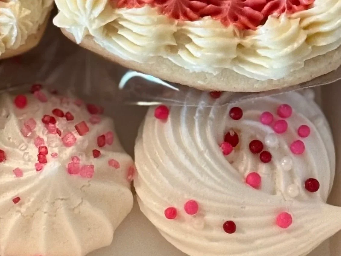 Close-up of three decorated cookies; the top cookie has pink and white frosting, and the bottom two are meringue cookies with pink sprinkles.